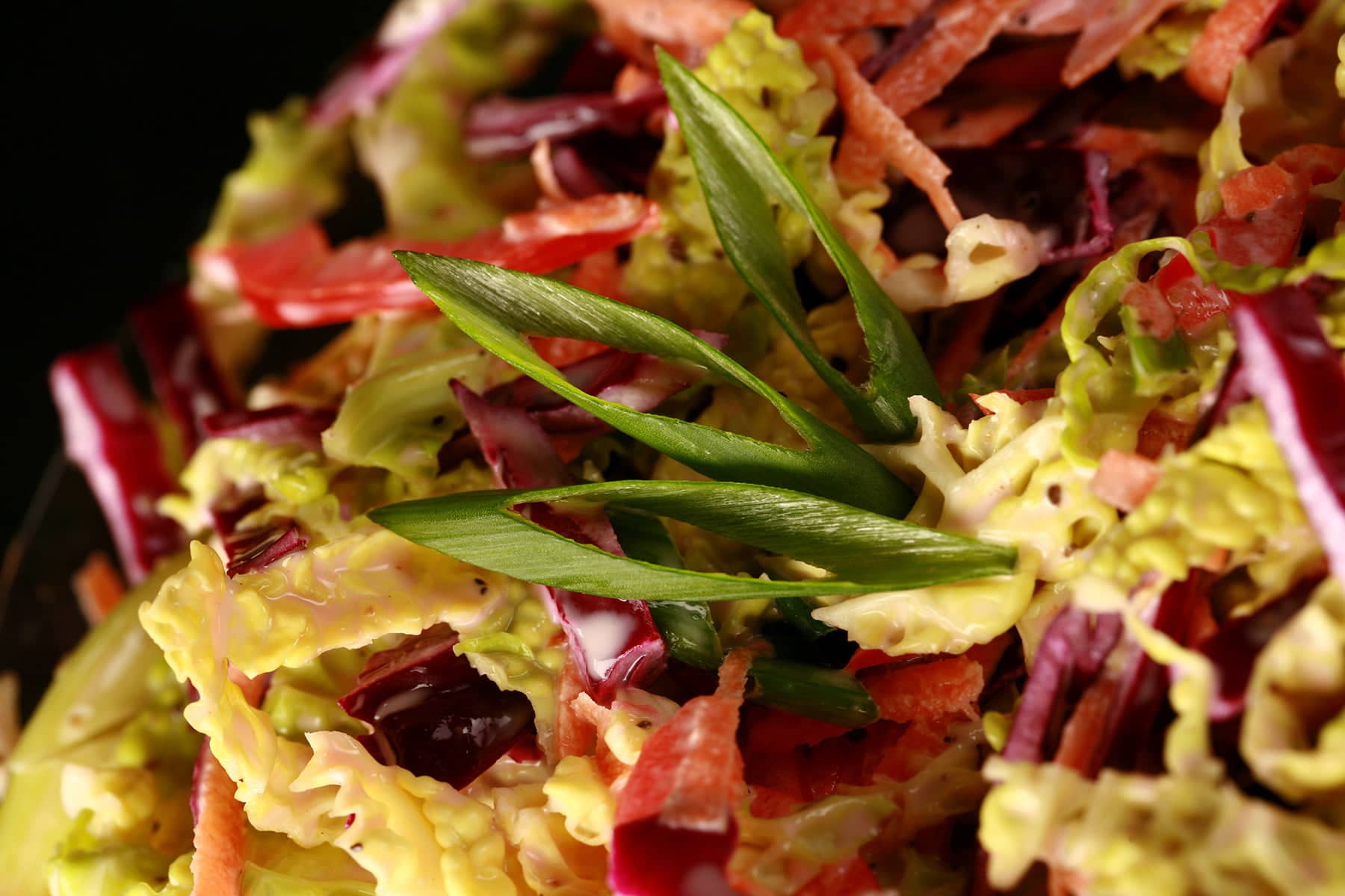 A glass bowl is filled with a colourful coleslaw. Red pepper, purple and green cabbage, carrot, and green onions are all visible.