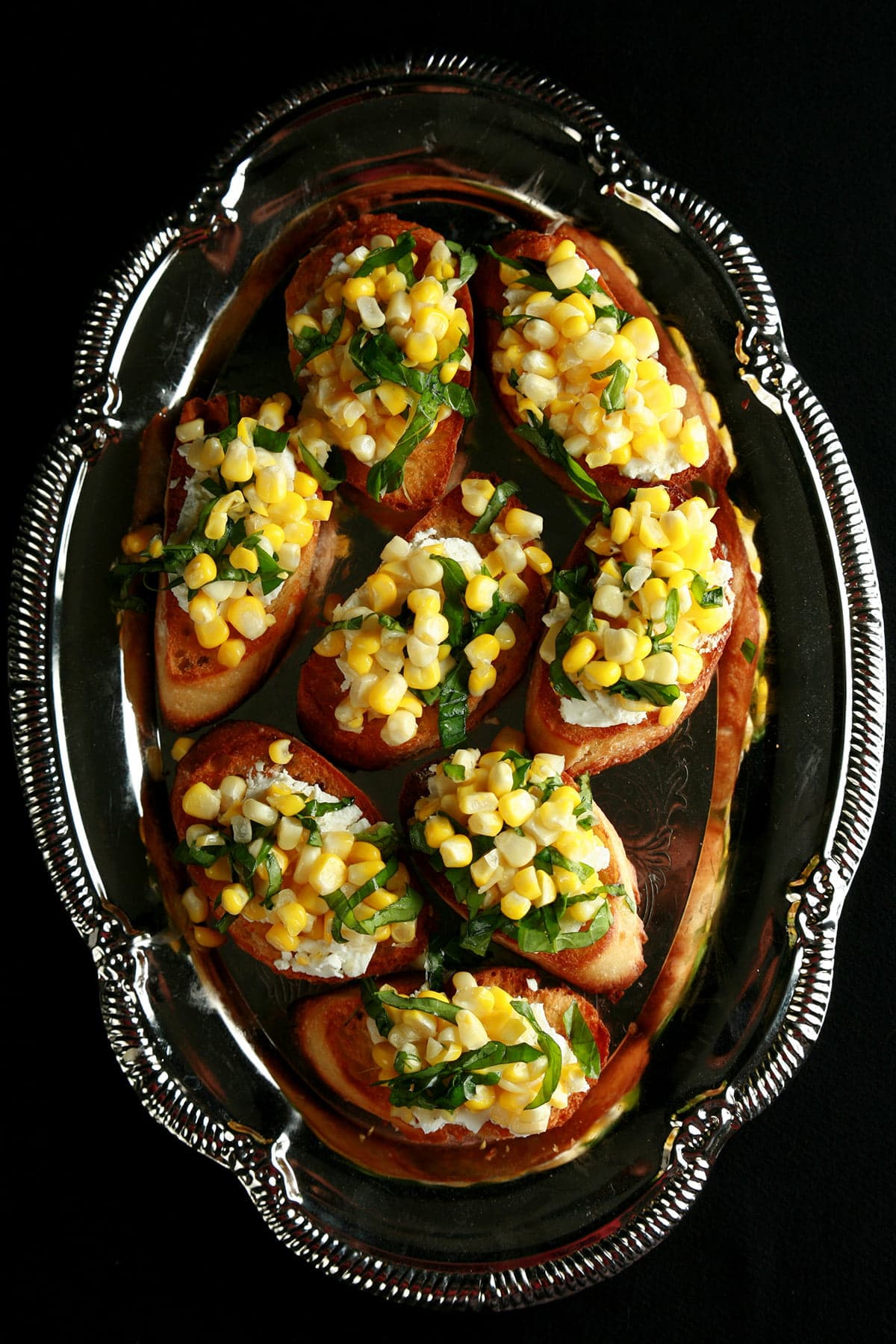 A silver, oval shaped platter, filled with sweet corn bruschetta - slices of toasted baguette, smeared with goat cheese, and topped with sweet corn kernels, basil, and balsamic vinegar.