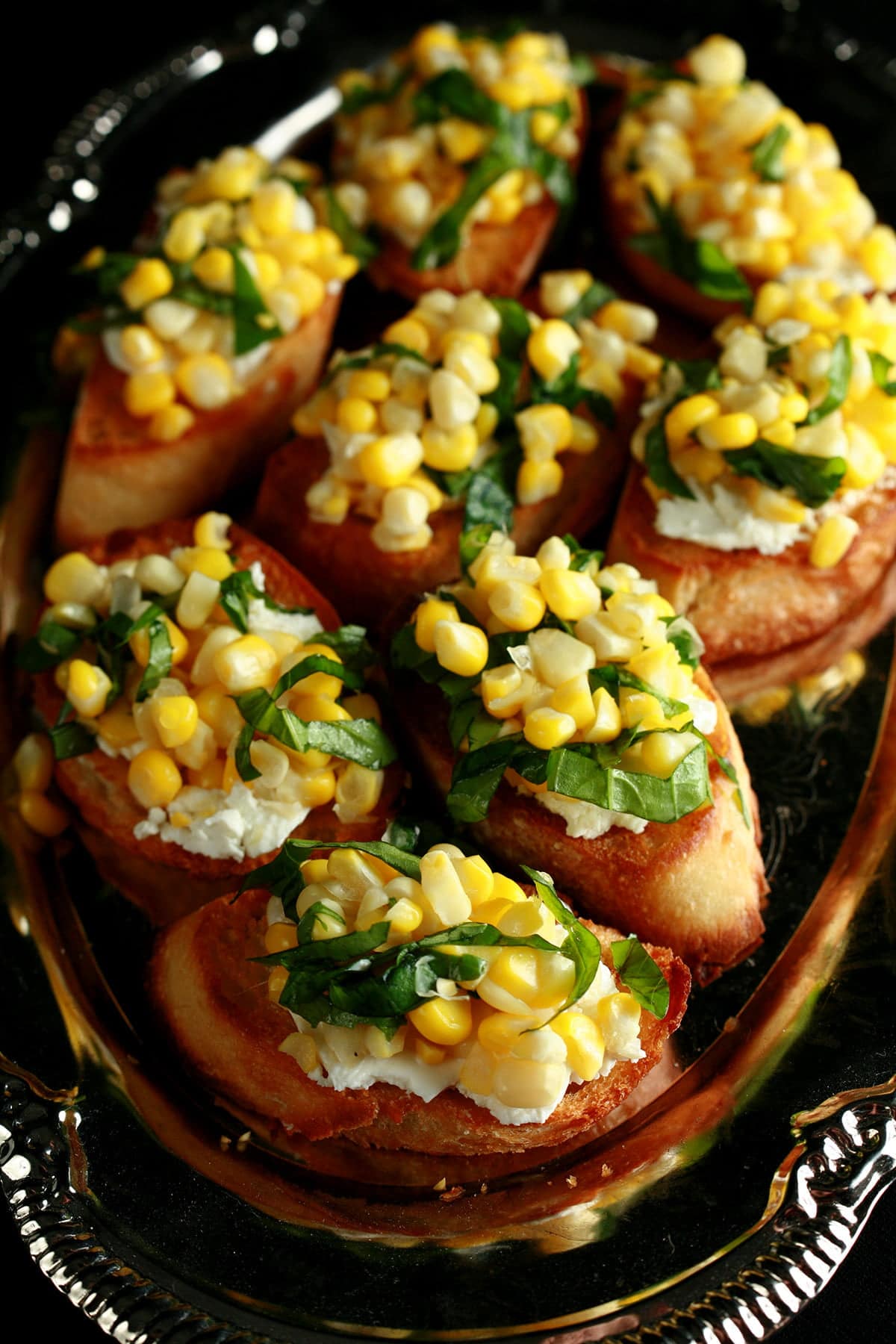 A silver, oval shaped platter, filled with sweet corn bruschetta - slices of toasted baguette, smeared with goat cheese, and topped with sweet corn kernels, basil, and balsamic vinegar.