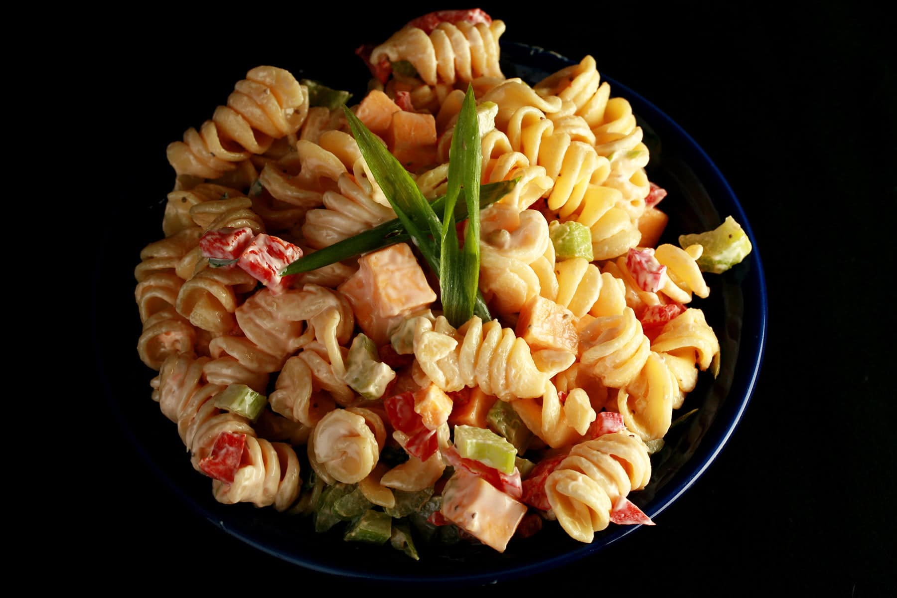 A blue bowl overflowing with a rotini based pasta salad. Cubes of cheese, red peppers, celery slices, and green onion are all visible.