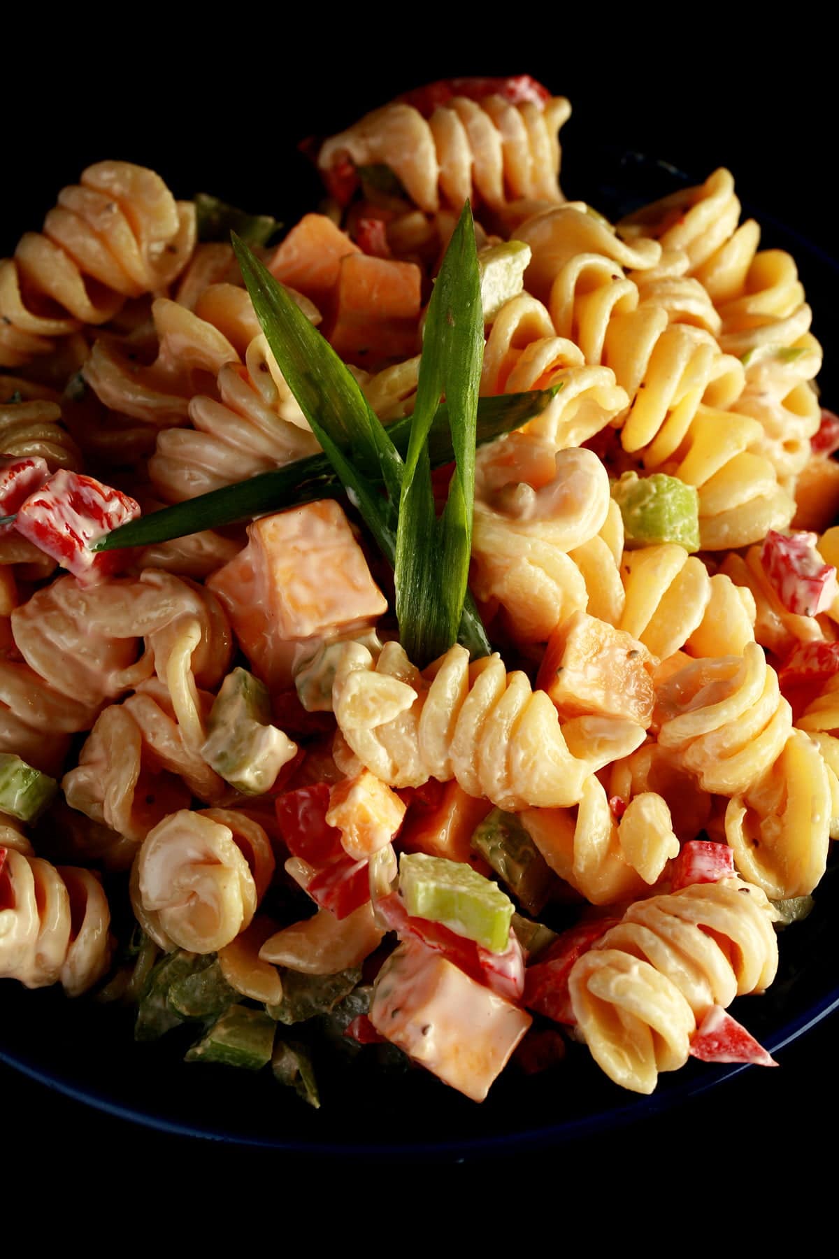 A blue bowl overflowing with a rotini based macaroni salad. Cubes of cheese, red peppers, celery slices, and green onion are all visible.