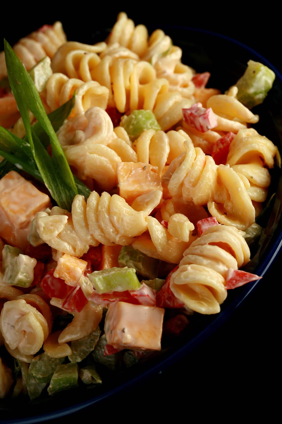 A blue bowl overflowing with a rotini based macaroni salad. Cubes of cheese, red peppers, celery slices, and green onion are all visible.