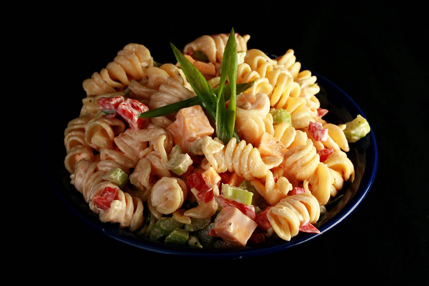 A blue bowl overflowing with a rotini based pasta salad. Cubes of cheese, red peppers, celery slices, and green onion are all visible.