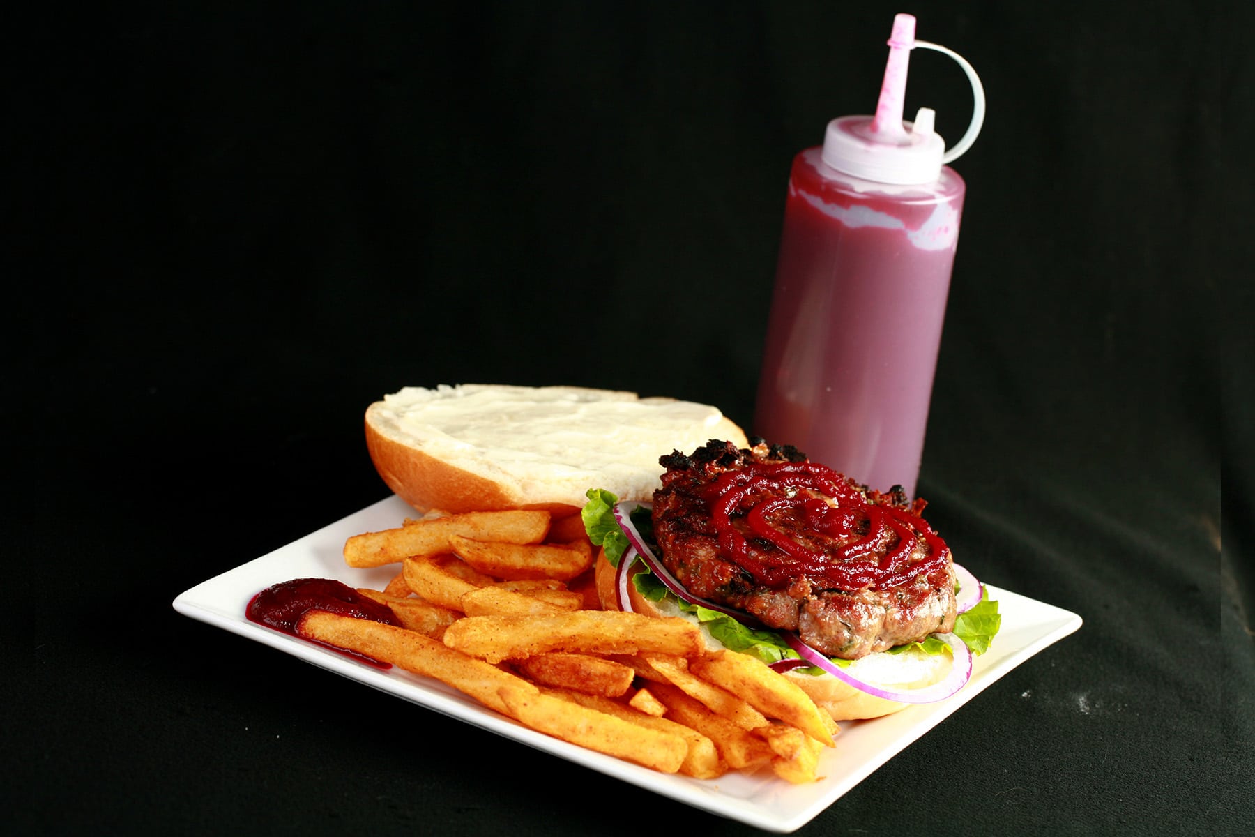An open faced burger on a plate with fries. The burger has a large ruby red swirl of beet ketchup on it, and there's a bottle of the same, behind the plate.