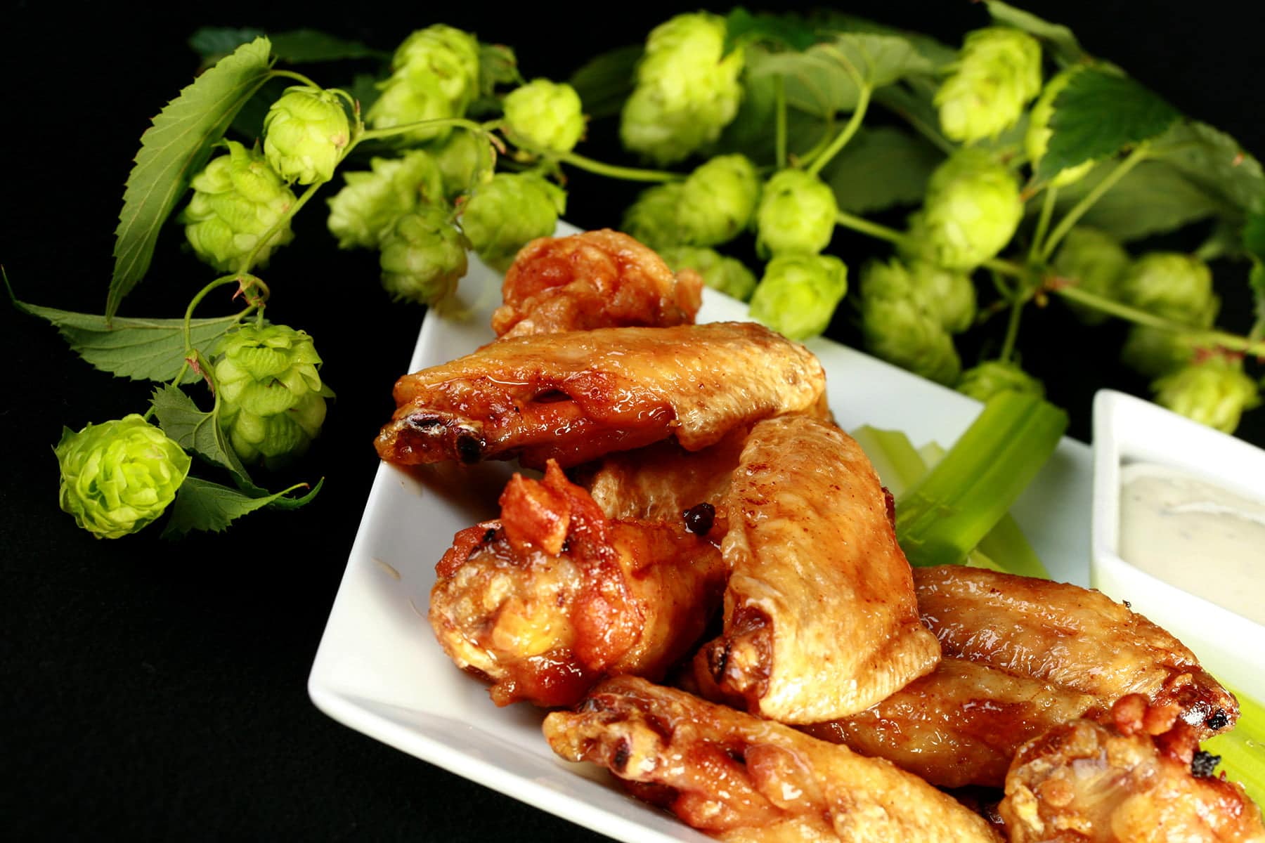 A plate of deep fried, Hoppy Citrus IPA Glazed Wings. They;re on a plate with celery sticks and a little bowl of ranch sauce, and the plate is surrounded by fresh hop bines.