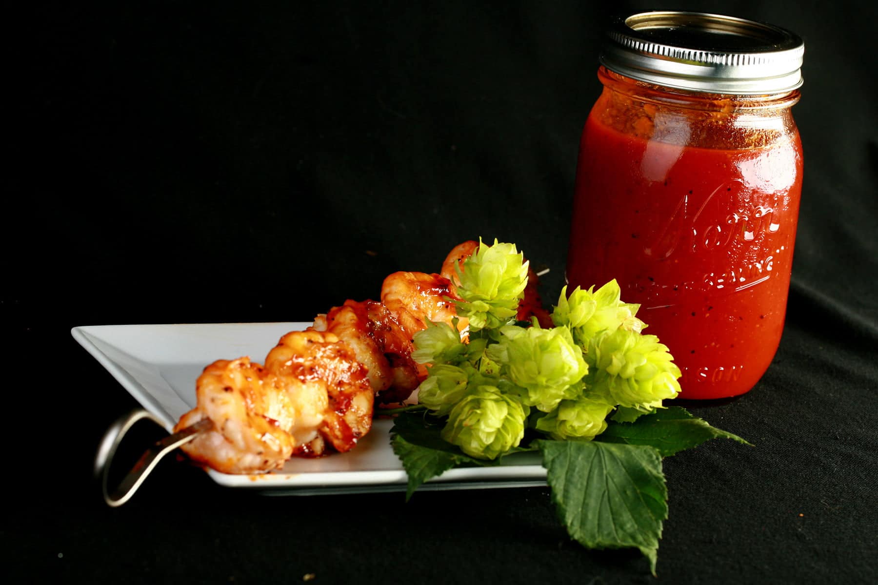 A jar of hoppy IPA BBQ Sauce, next to some fresh hops, and a plate with a skewer of grilled shrimp on it.