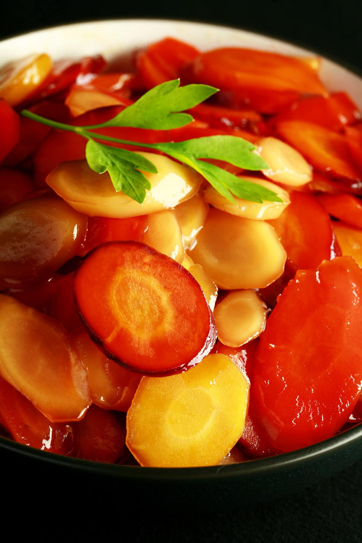 A bowl of brightly coloured carrot slices, in a shiny glaze. There is a small piece of flat leaf parsley on top, as garnish.