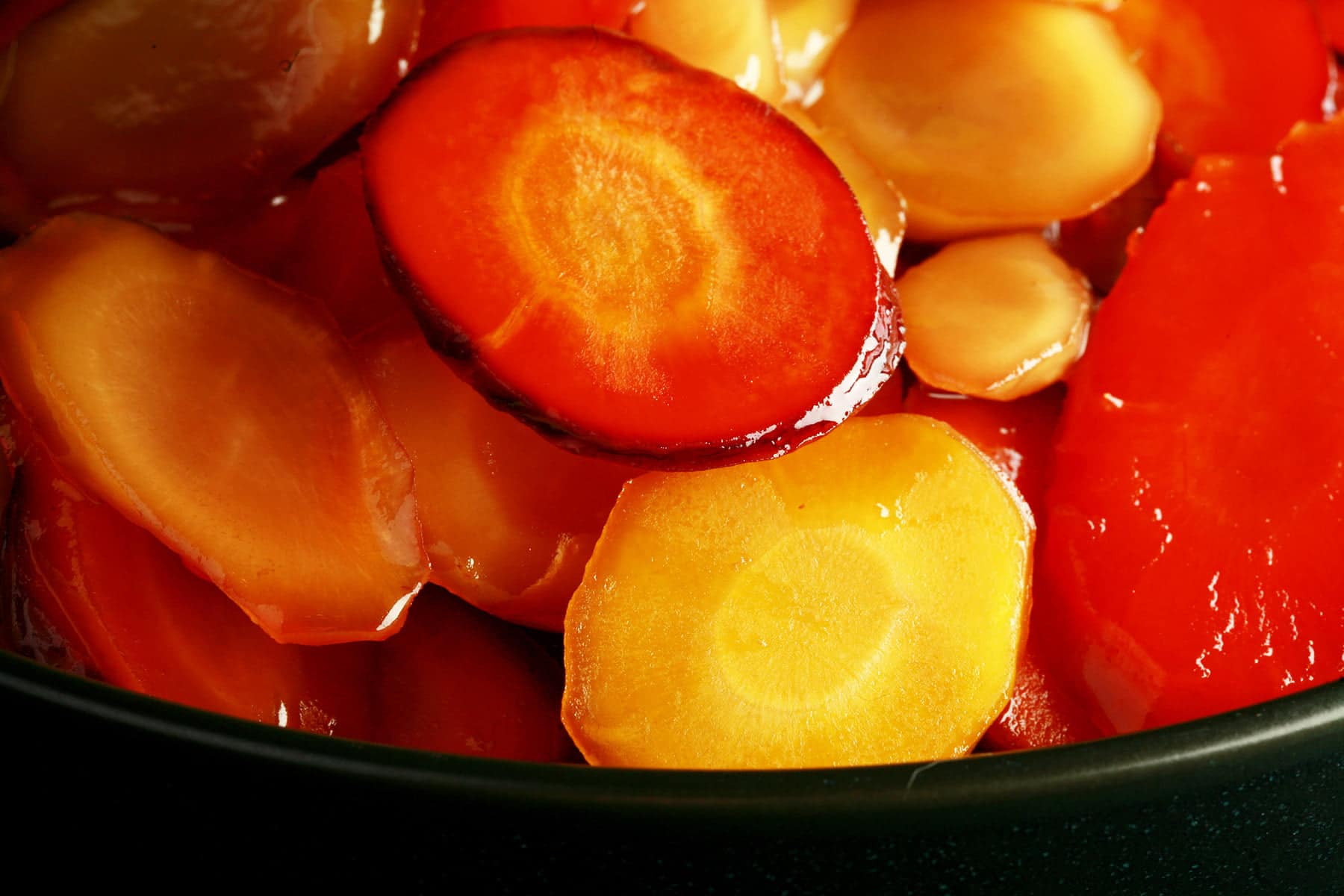 A bowl of brightly coloured carrot slices, in a shiny glaze. There is a small piece of flat leaf parsley on top, as garnish.