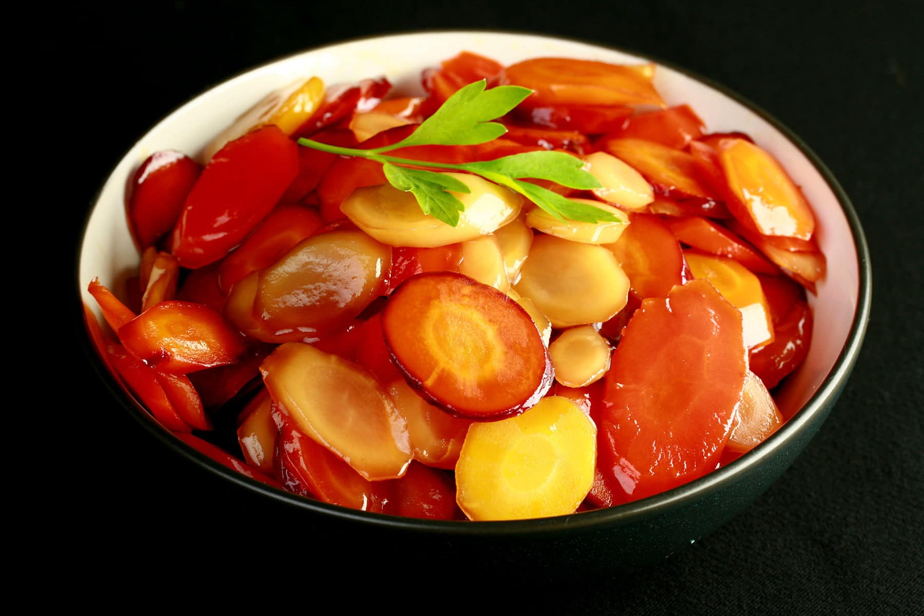 A bowl of brightly coloured carrot slices, in a shiny glaze. There is a small piece of flat leaf parsley on top, as garnish.