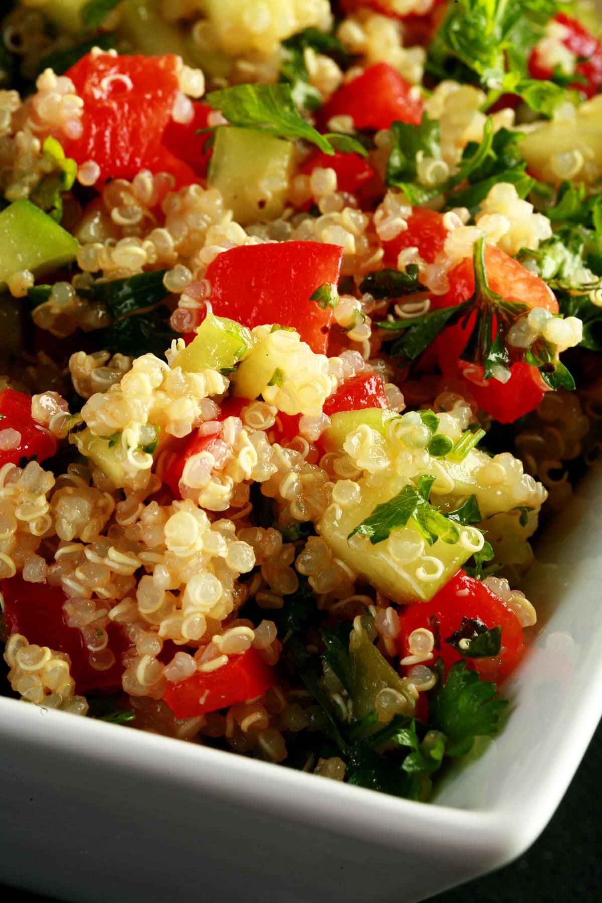 A square, white bowl holds a no-tomato quinoa based tabbouleh.  Beige quinoa, red pepper, cucumber, and parsley is visible throughout.