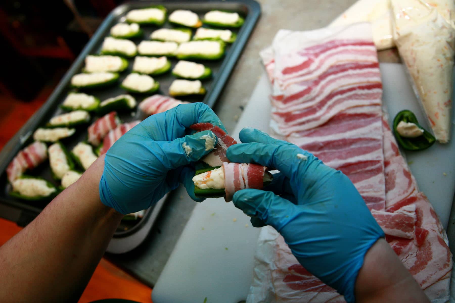 Gloved hands wrap a cheese stuffed jalapeno half with bacon.