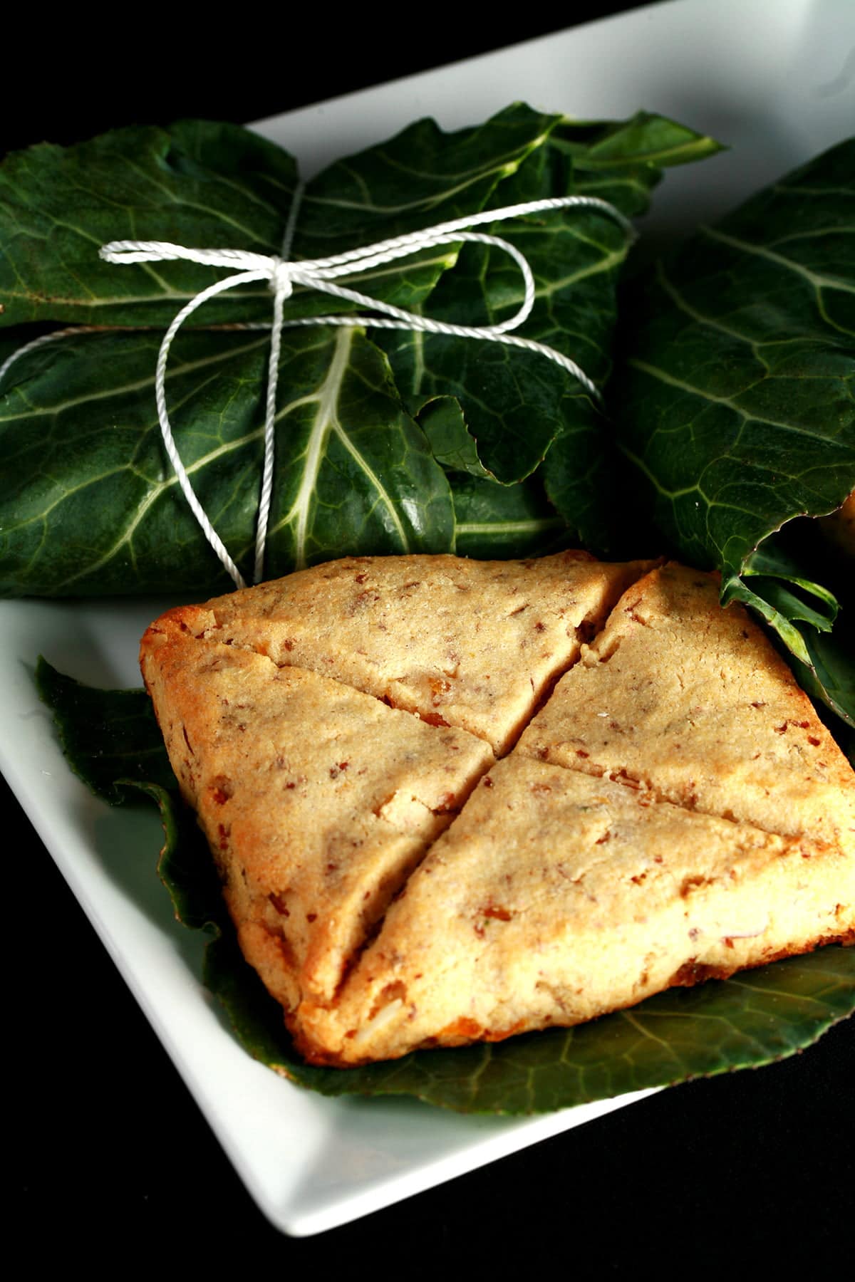 A square white plate displays 3 pieces of Lembas bread. Two of the square biscuits - each marked with an X cut - are unwrapped, while the 3 is still neatly packaged in a large leaf, and tied with twine.