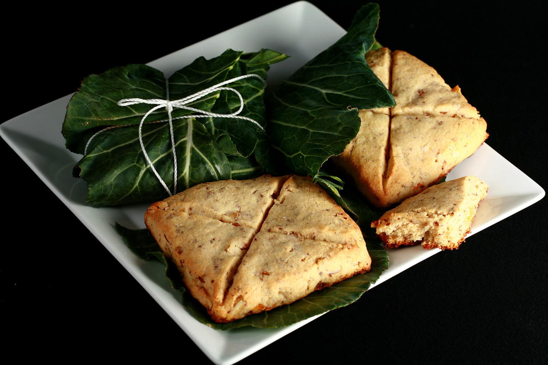 A square white plate displays 3 pieces of Lembas bread. Two of the square biscuits - each marked with an X cut - are unwrapped, while the 3 is still neatly packaged in a large leaf, and tied with twine.
