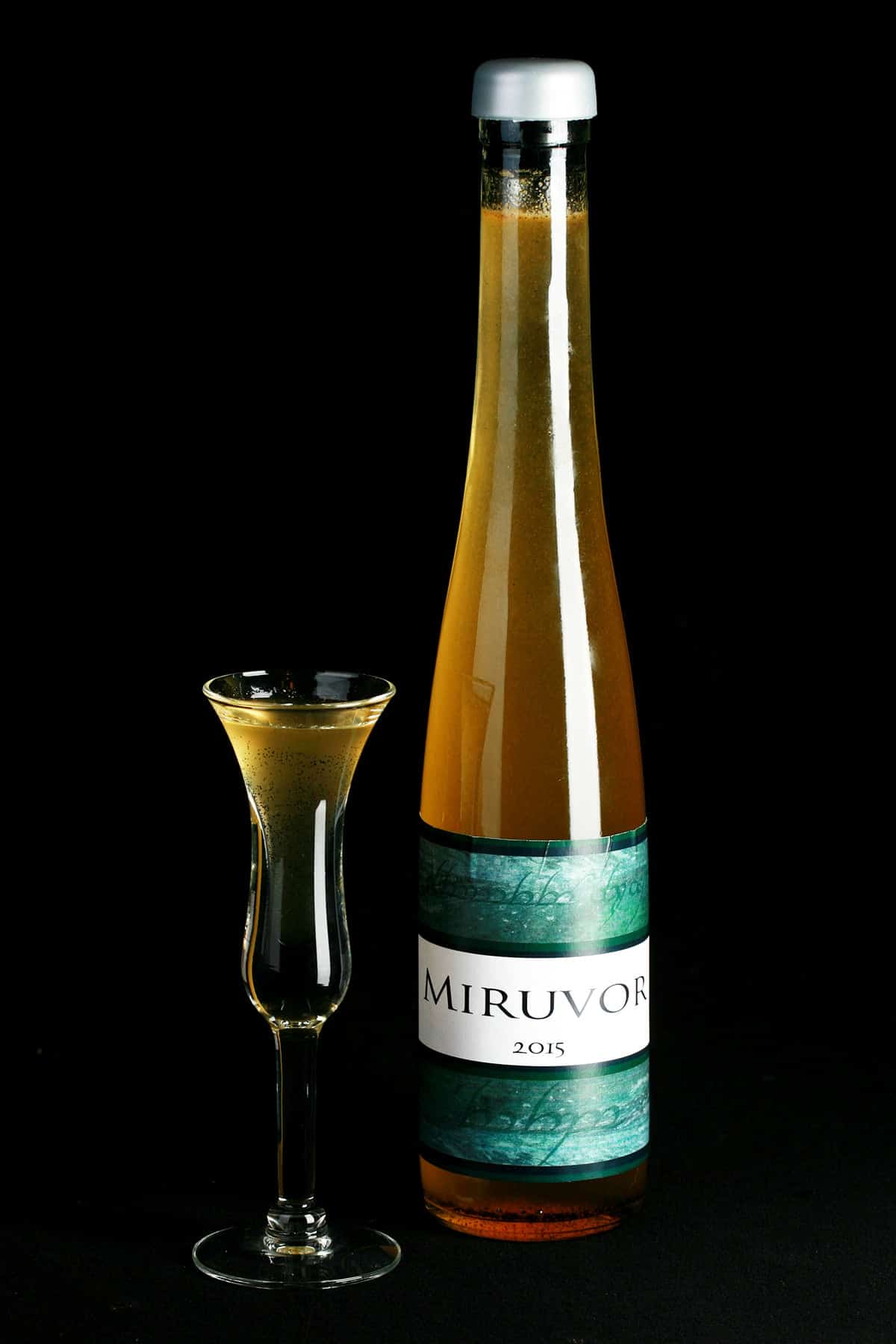 A tall, slender wine bottle filled with a straw coloured liqueur is pictured next to a fluted shot glass filled with the same.