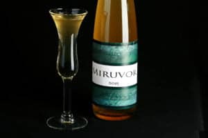A tall, slender wine bottle filled with a straw coloured liqueur is pictured next to a fluted shot glass filled with the same. The bottle is labelled "Miruvor".