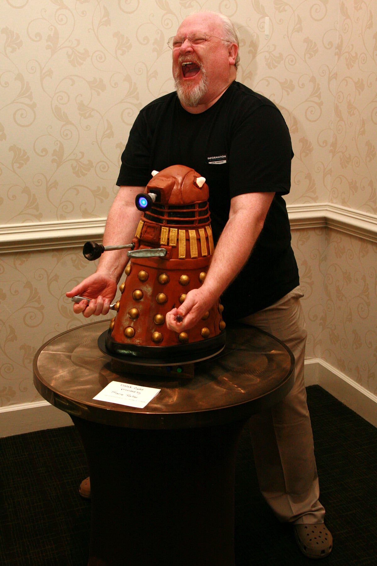 Colin Baker jams 2 serving knives into the Dalek Cake while posing.