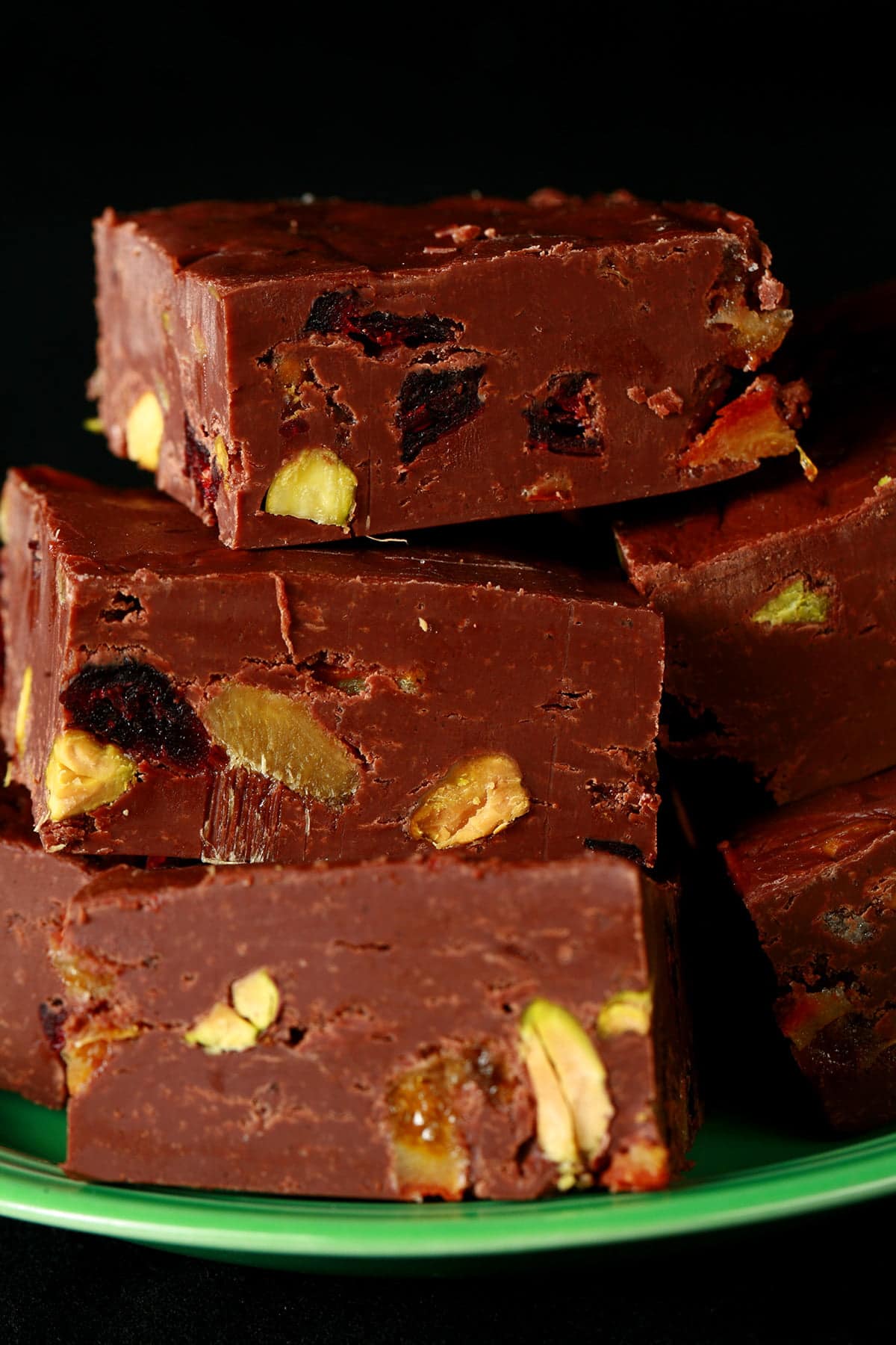 A green plate with a pile of chocolate fudge on it. Each square of fudge is studded with nuts, dried cranberries, candied orange peel, and candied ginger.