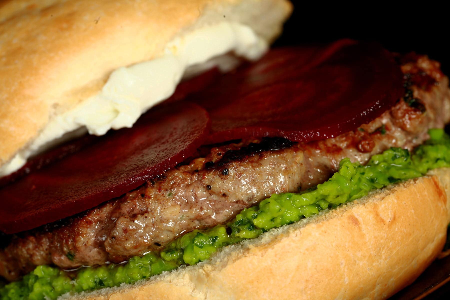 Close up view of a lamb burger with a green spread, a goat cheese spread, and beet slices on it.