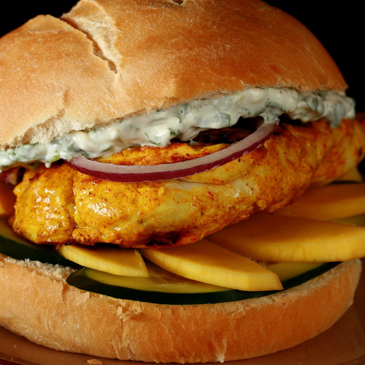 Tandoori Marinated chicken breast with cucumbers, mango, red onions, and a cilantro mint mayo, on a bun.