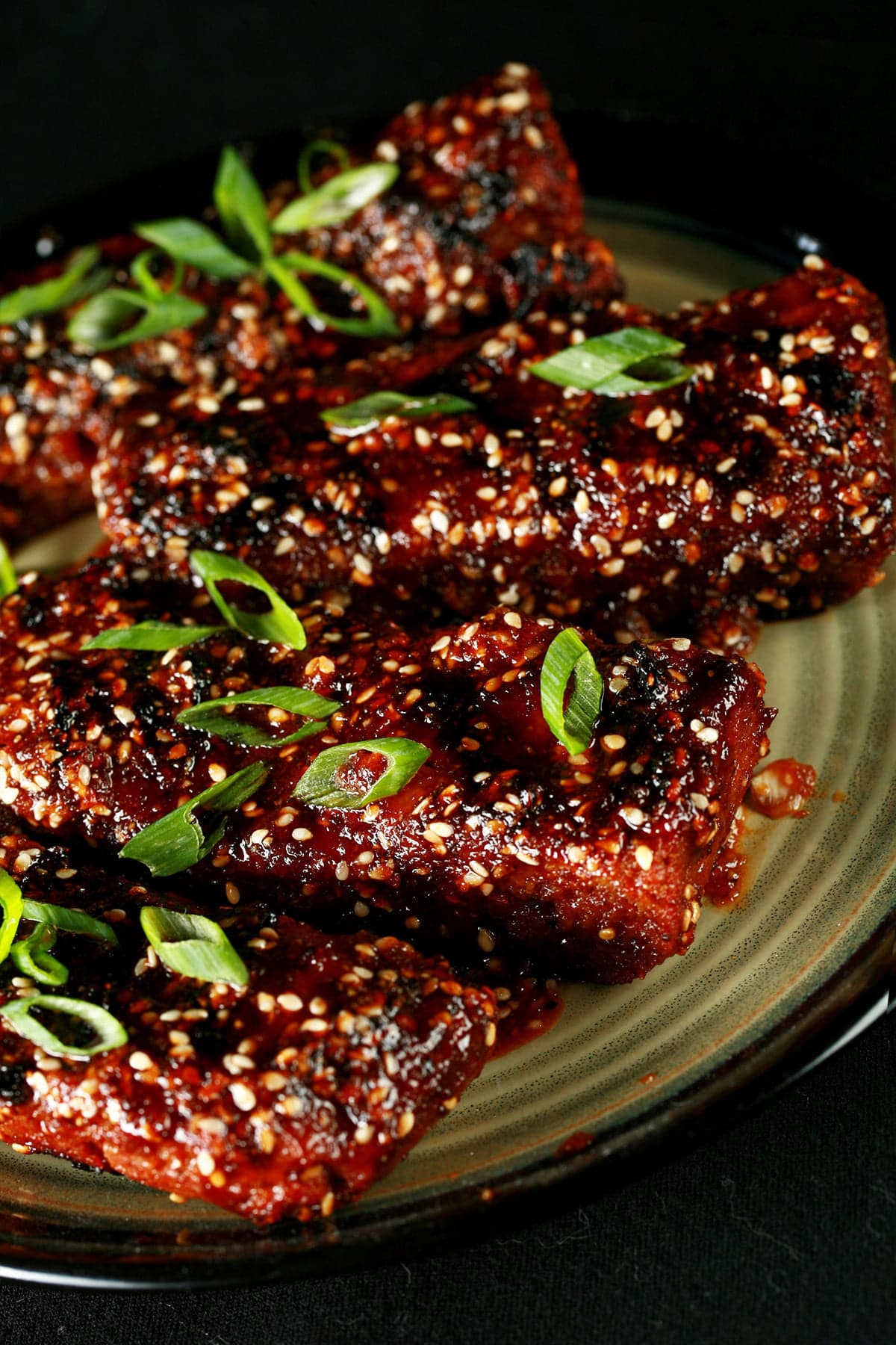 Close up photo of a plate of vegan boneless ribs. They're covered in a red-brown sauce, sesame seeds, and sliced green onion.