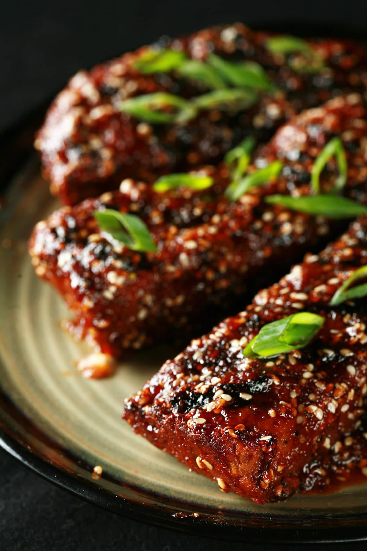 Close up photo of a plate of vegetarian boneless ribs. They're covered in a red-brown sauce, sesame seeds, and sliced green onion.