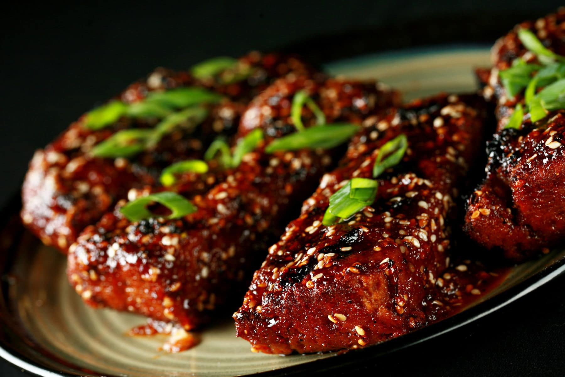 Close up photo of a plate of vegetarian boneless ribs. They're covered in a red-brown sauce, sesame seeds, and sliced green onion.