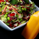 A close up view of rainbow salad. Cucumber slices, edamame, red peppers, carrot, purple cabbage, and red onions are all visible. The salad is in a large white bowl, with a bottle of a bright orange carrot ginger dressing next to it.