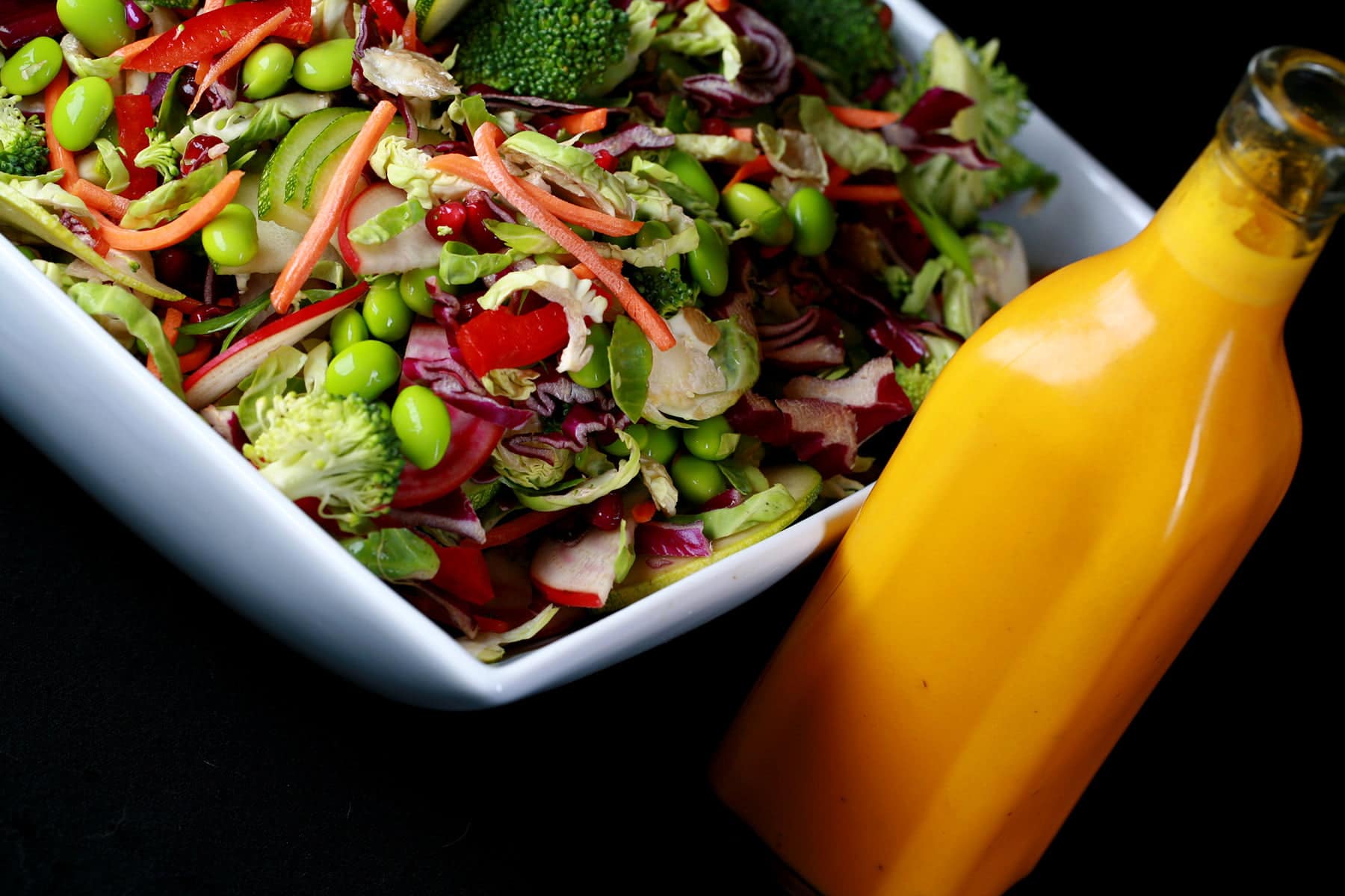 A close up view of rainbow salad. Cucumber slices, edamame, red peppers, carrot, purple cabbage, and red onions are all visible.  The salad is in a large white bowl, with a bottle of a bright orange carrot ginger dressing next to it.
