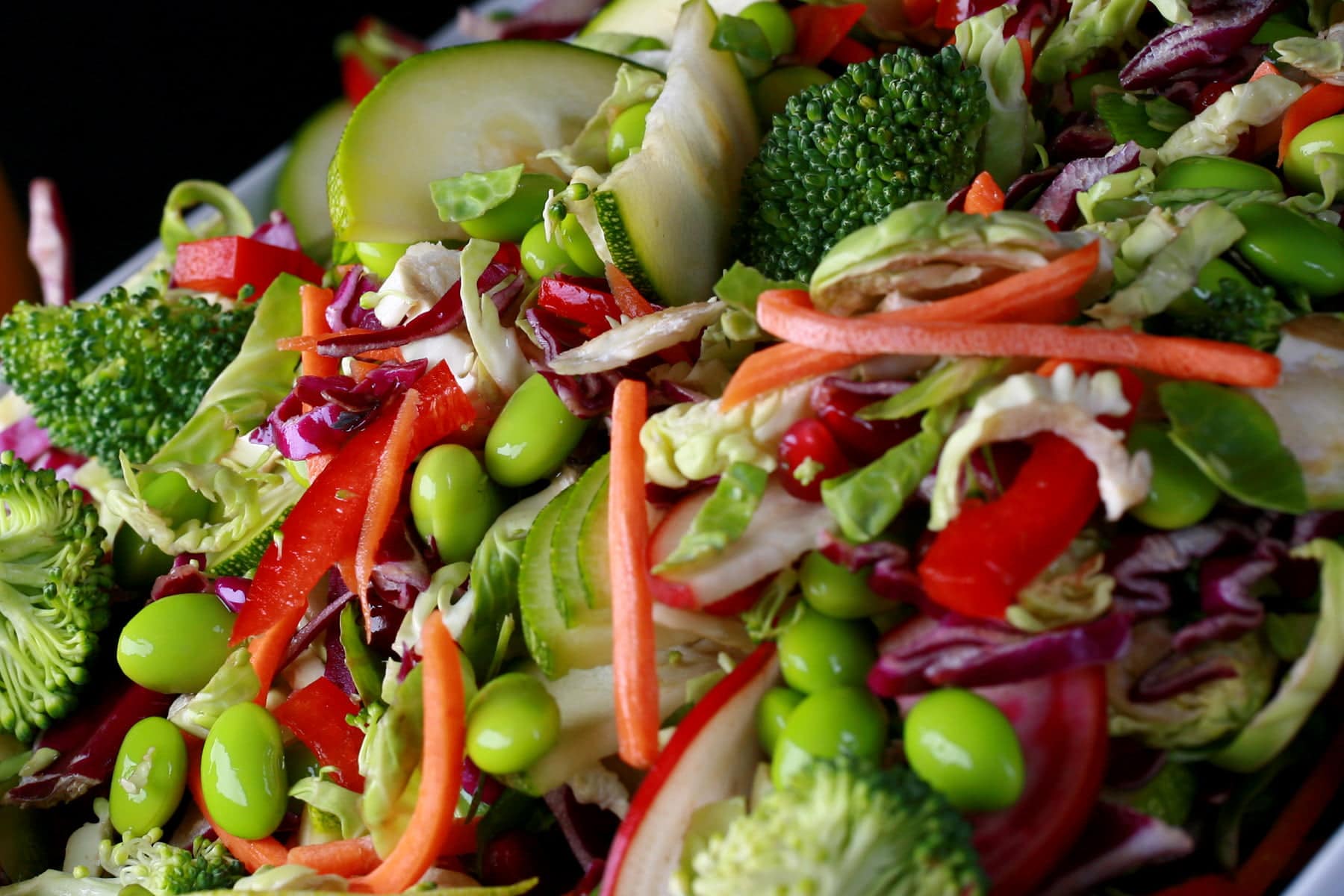 A close up view of rainbow salad. Cucumber slices, edamame, red peppers, carrot, purple cabbage, and red onions are all visible.