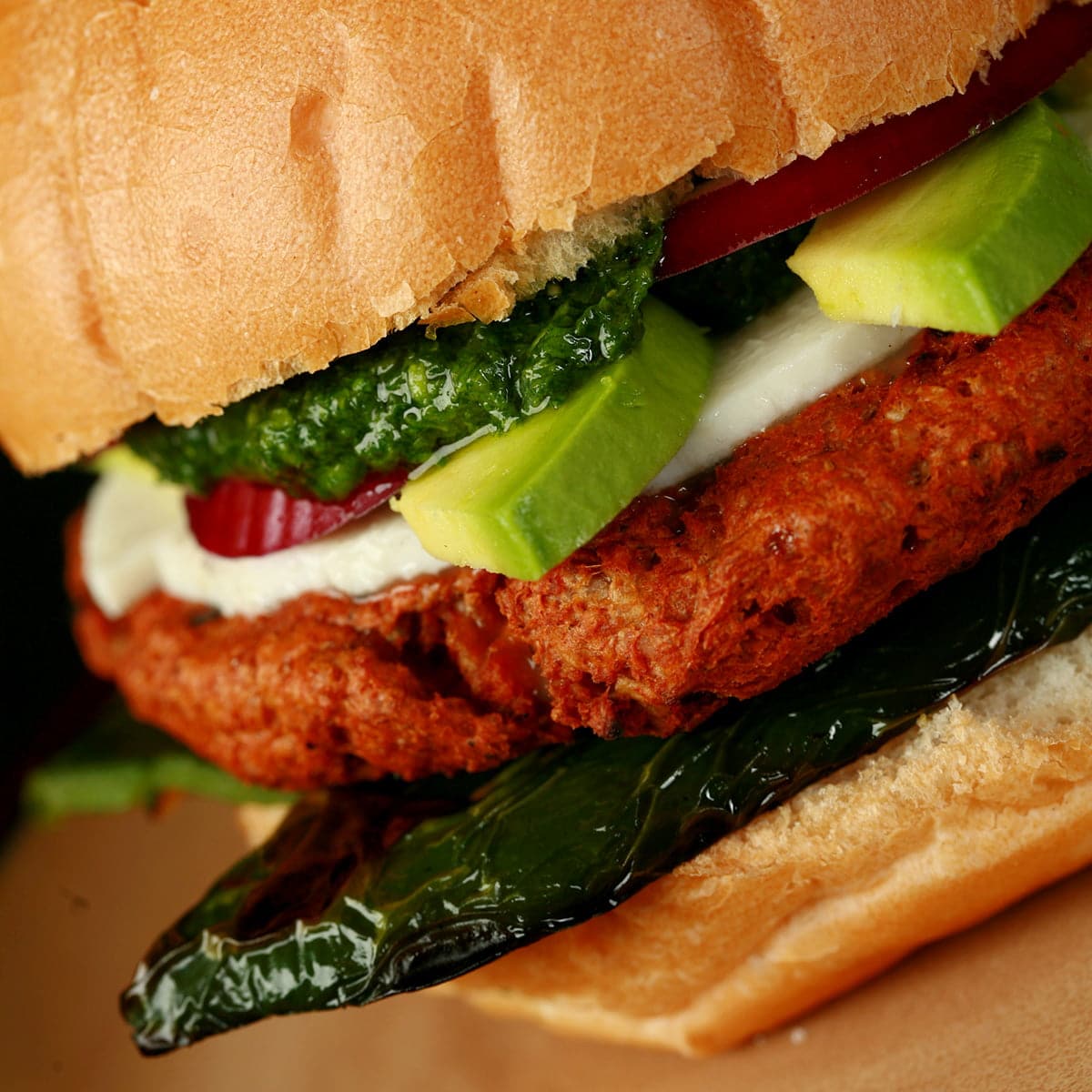 A close up view of a fully decked out vegetarian chorizo burger. Aside from the patty, a slice of roasted poblano, slices of red onion, a white cheese, and avocados are seen. A deep green cilantro pesto tops it all off.