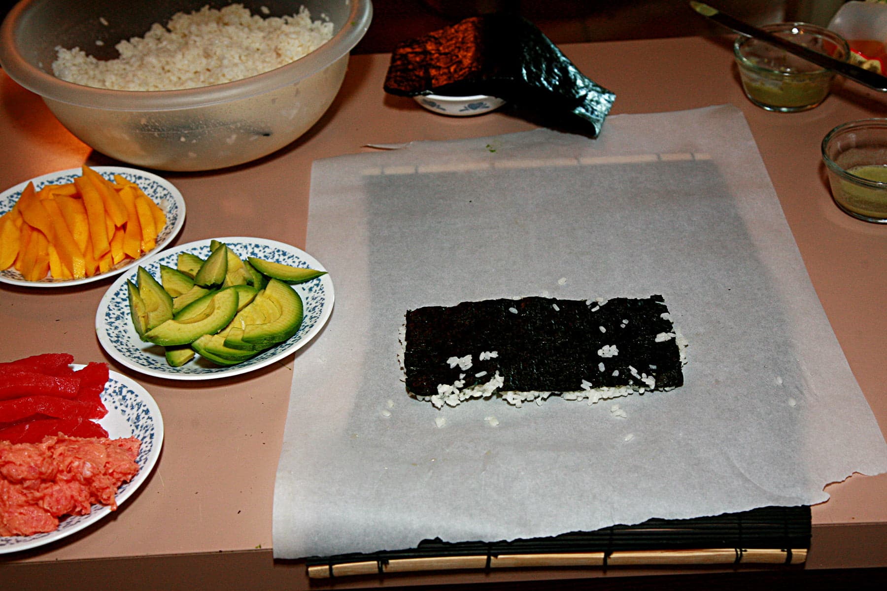 A sushi rolling station. A piece of nori with rice is laid out on a piece of parchment paper, with various sushi fillings laid out on plates next to it.