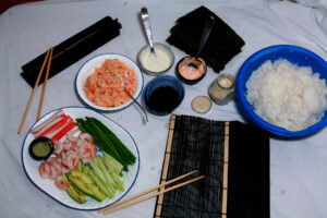 Various sushi fixings are shown laid out on a paper-lined table. There are plates of fillings, a bowl of rice, a stack of nori seets, and wrapping utensils.