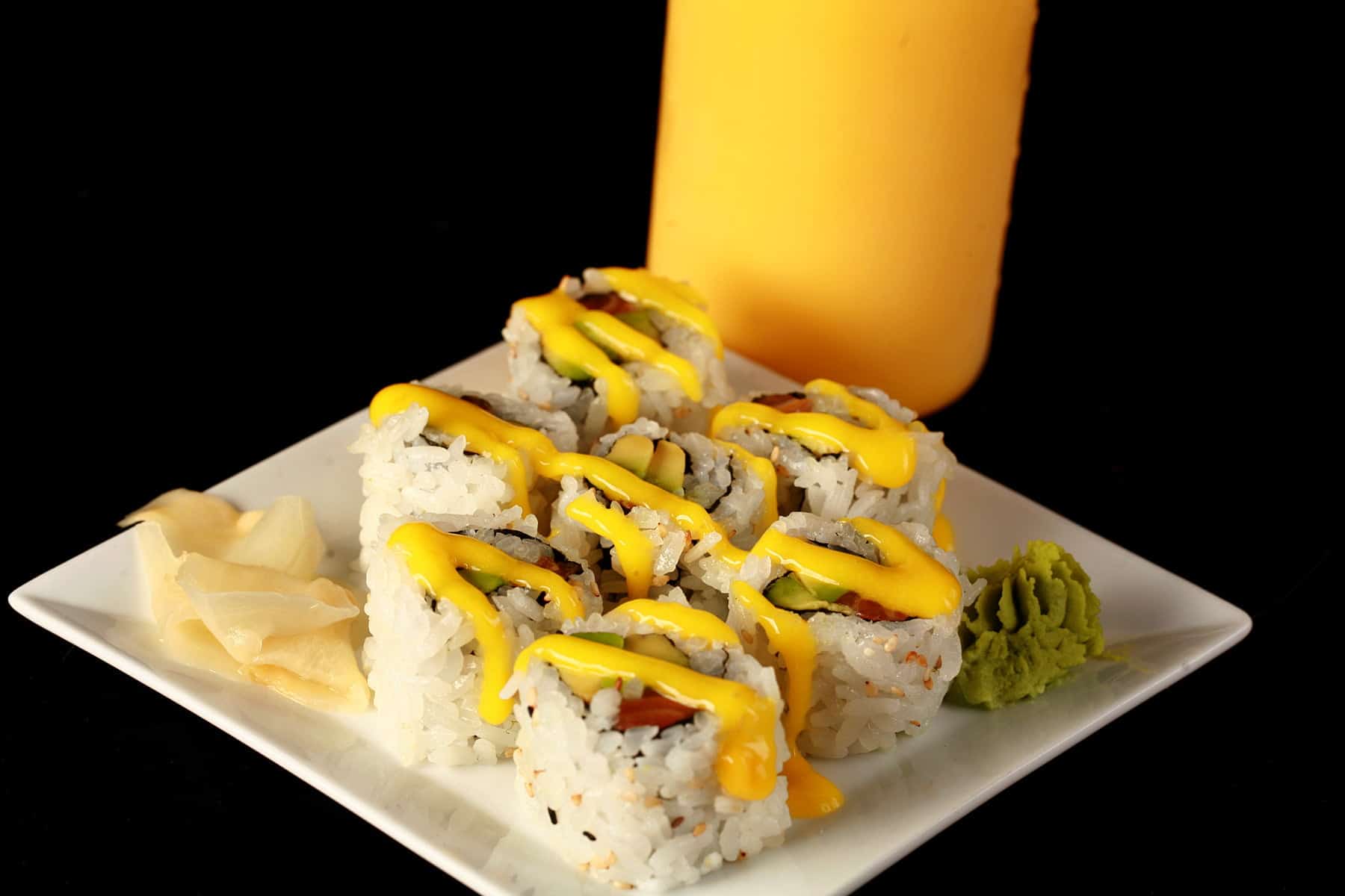 Close up image of a small plate of sushi, with yellow sauce drizzled across it. There is a bottle of yellow sauce - Mango Sauce for Sushi - behind the plate. This is one of 3 Sushi Sauce Recipes on this post.