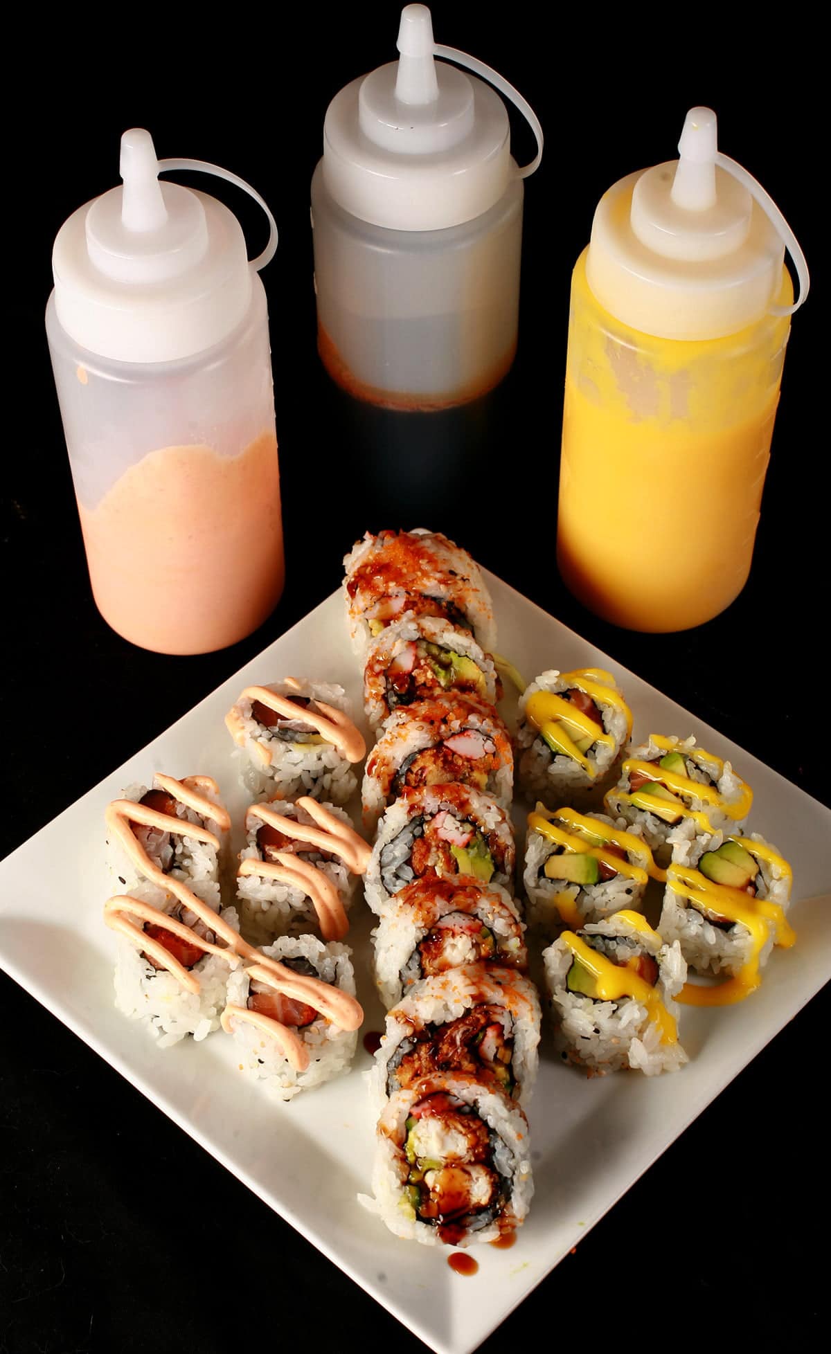 Close up image of 3 bottles of sauce - one yellow, one brown, and one pink. In front of the bottles is a plate of sushi, with the 3 colours of sauce drizzled across the sushi in squiggles