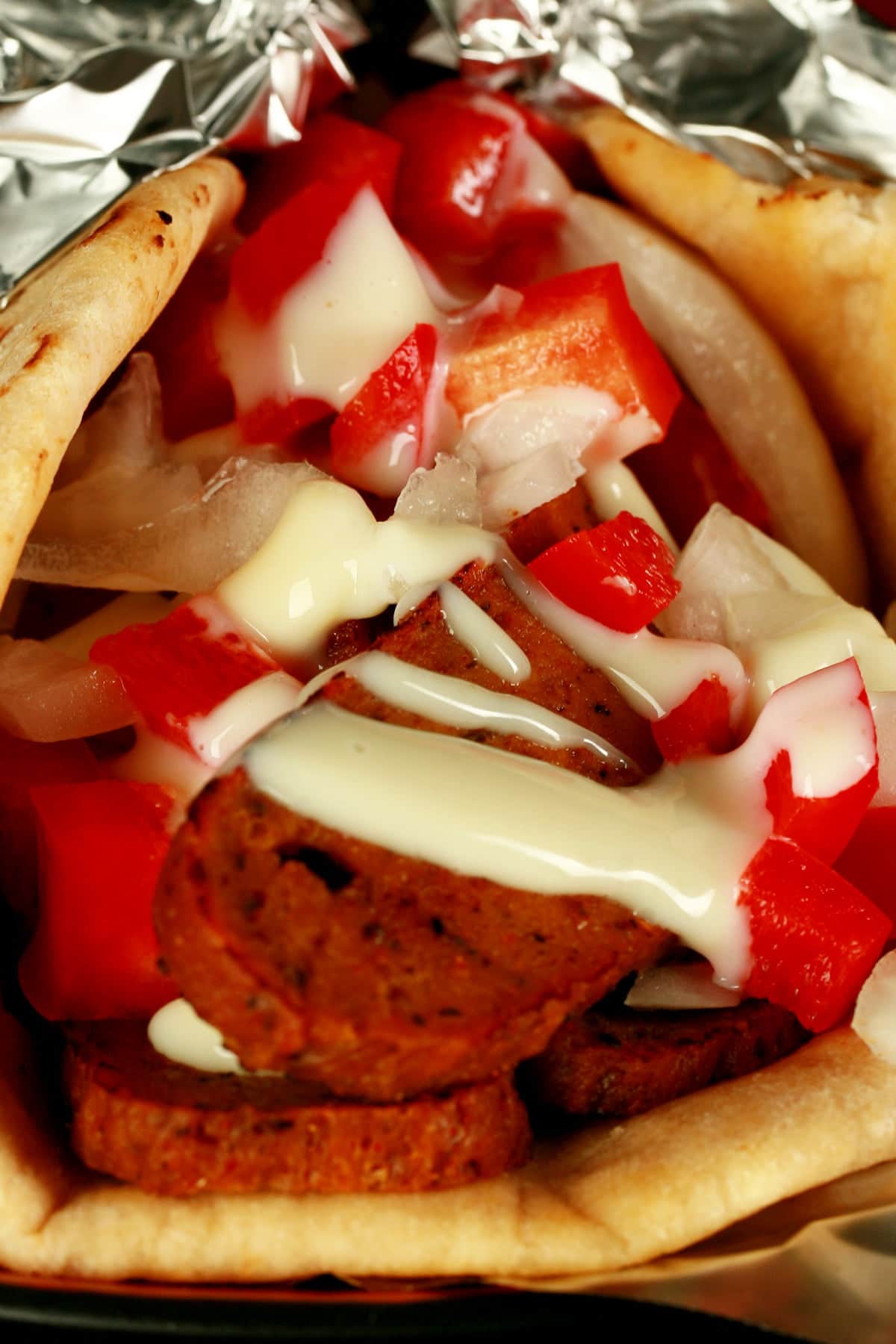 Close up view of a vegan donair - a pita bread folded around a pile of vegan donair "meat" slices, chopped red peppers, and donair sauce drizzled on top.