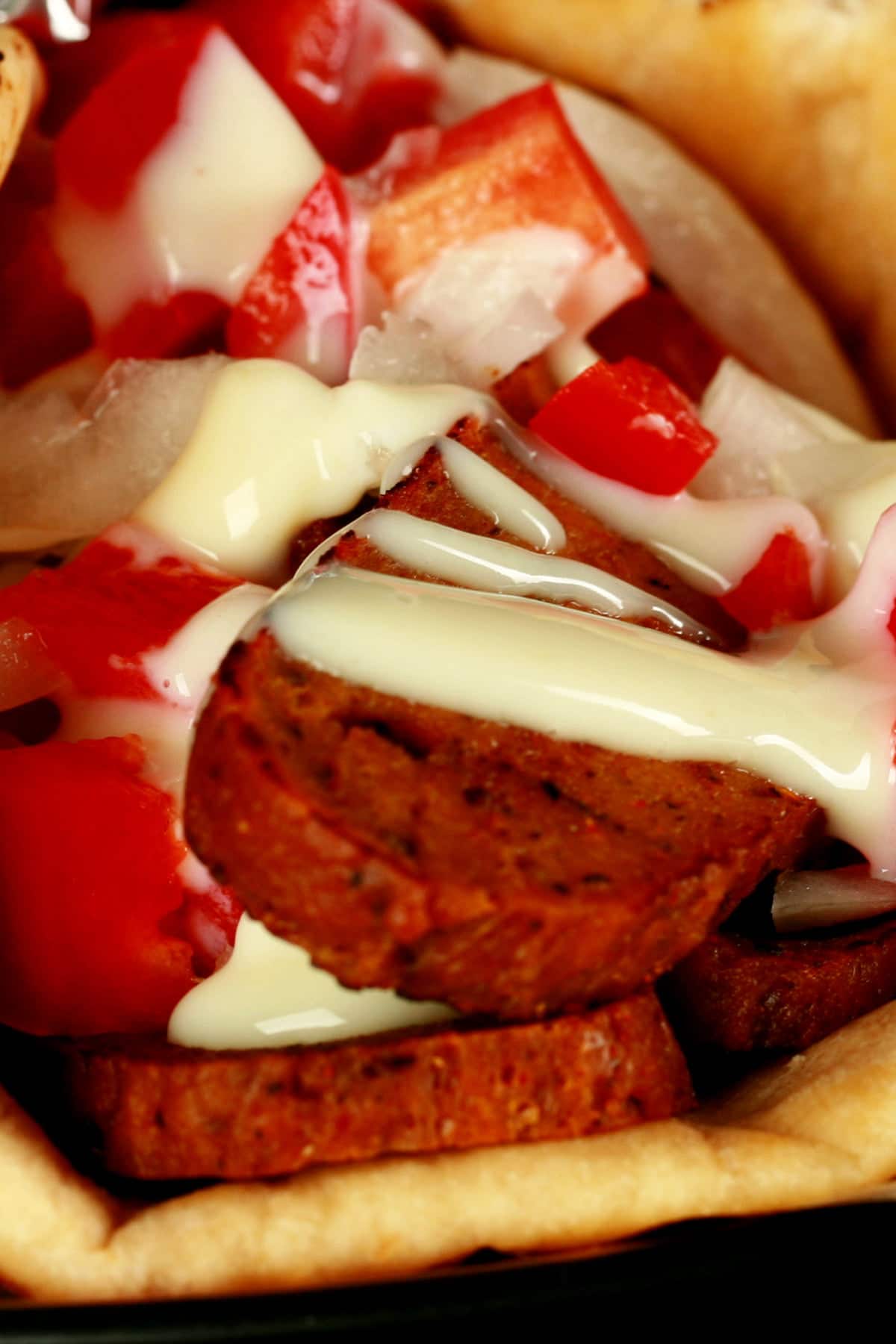 Close up view of a vegan donair - a pita bread folded around a pile of vegan donair "meat" slices, chopped red peppers, and donair sauce drizzled on top.