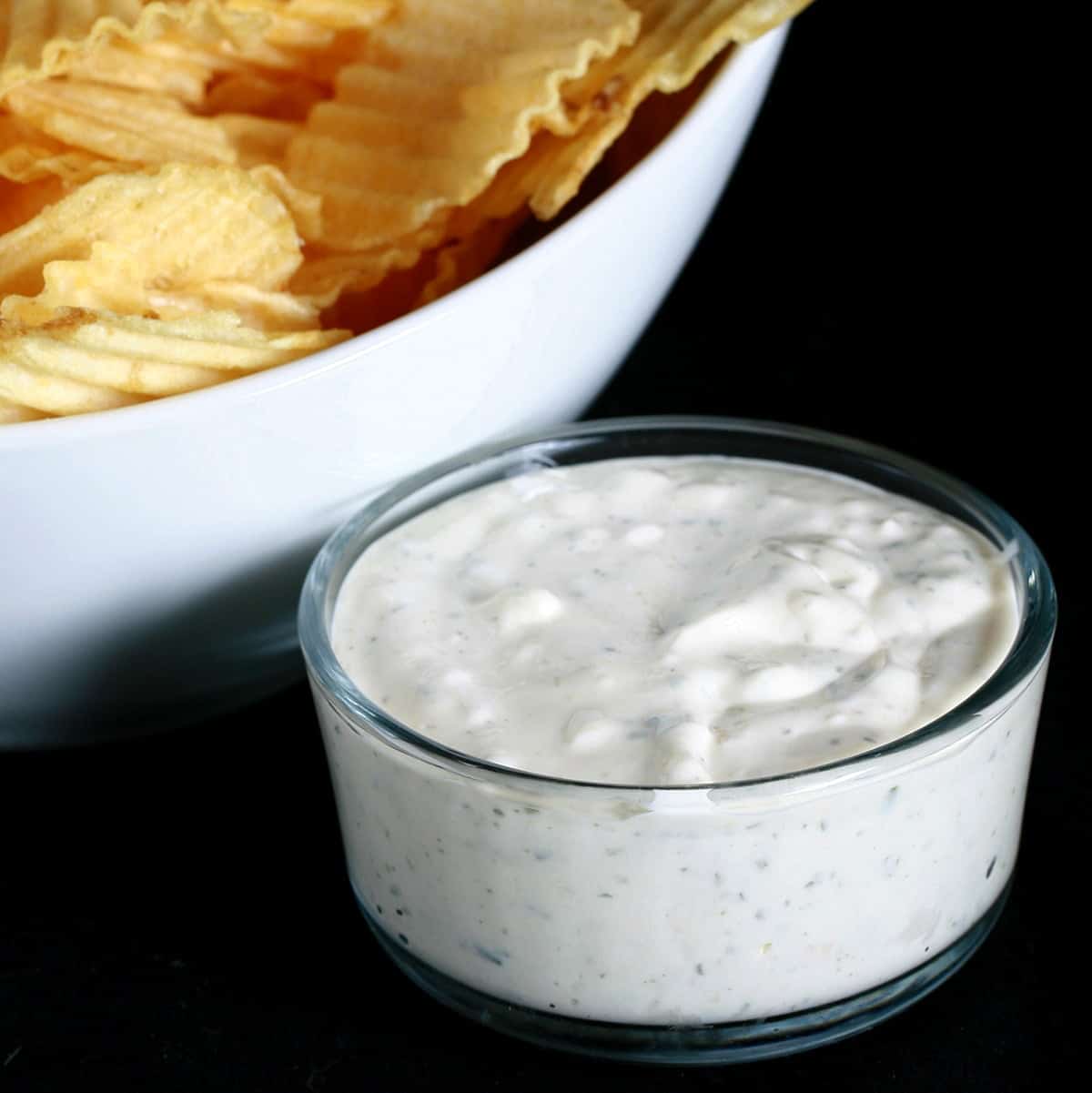 A close up view of a bowl of dill pickle cream cheese dip, next to a white bowl full of ripple chips.
