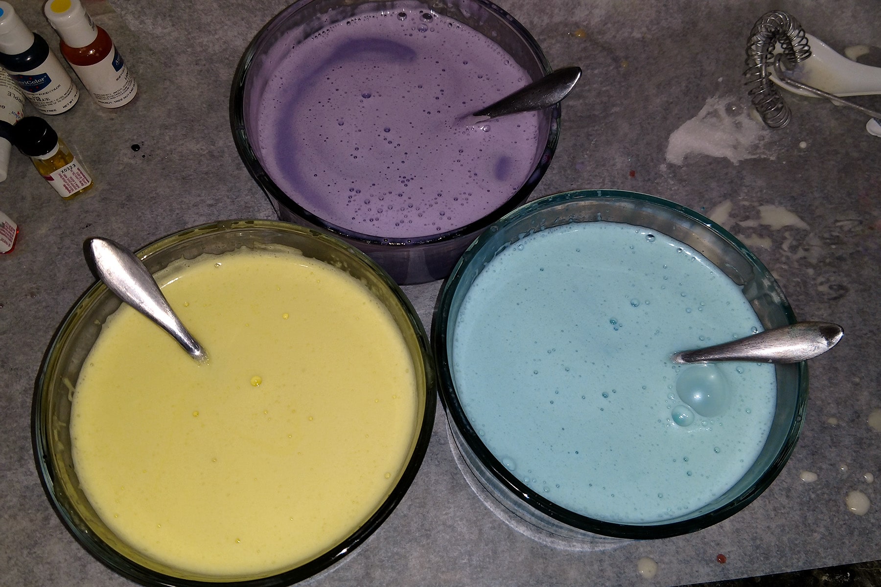 2 small glass mixing bowls with milky liquid in each. One is yellow, another is sky blue, and the last is purple.