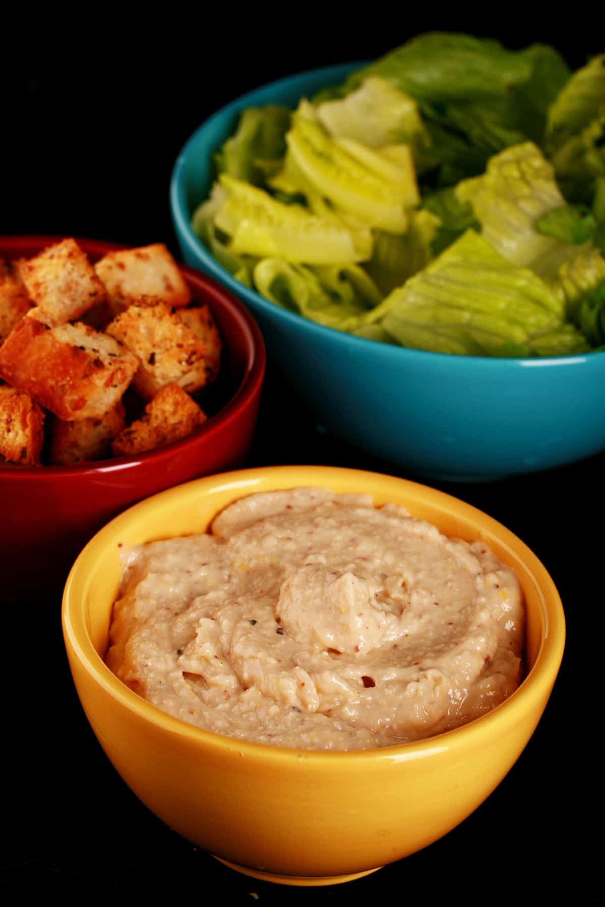 A large blue bowl full of chopped romaine lettuce, a yellow bowl of "extreme" Caesar salad, and a red bowl full of homemade croutons.