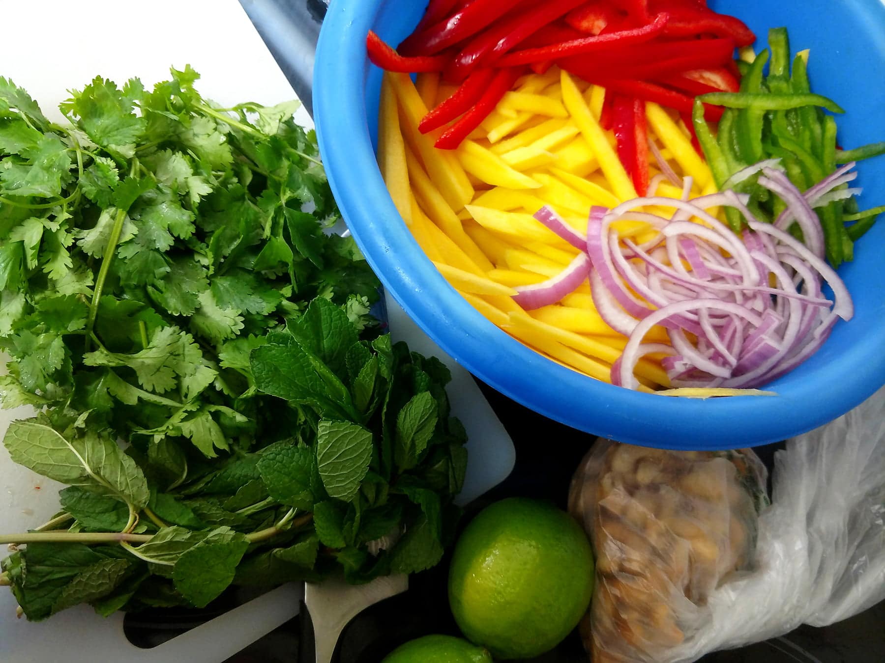 A blue mixing bowl holds slices of mango, red and green peppers, and red onions. Cilantro and mint is shown on a cutting board beside it.