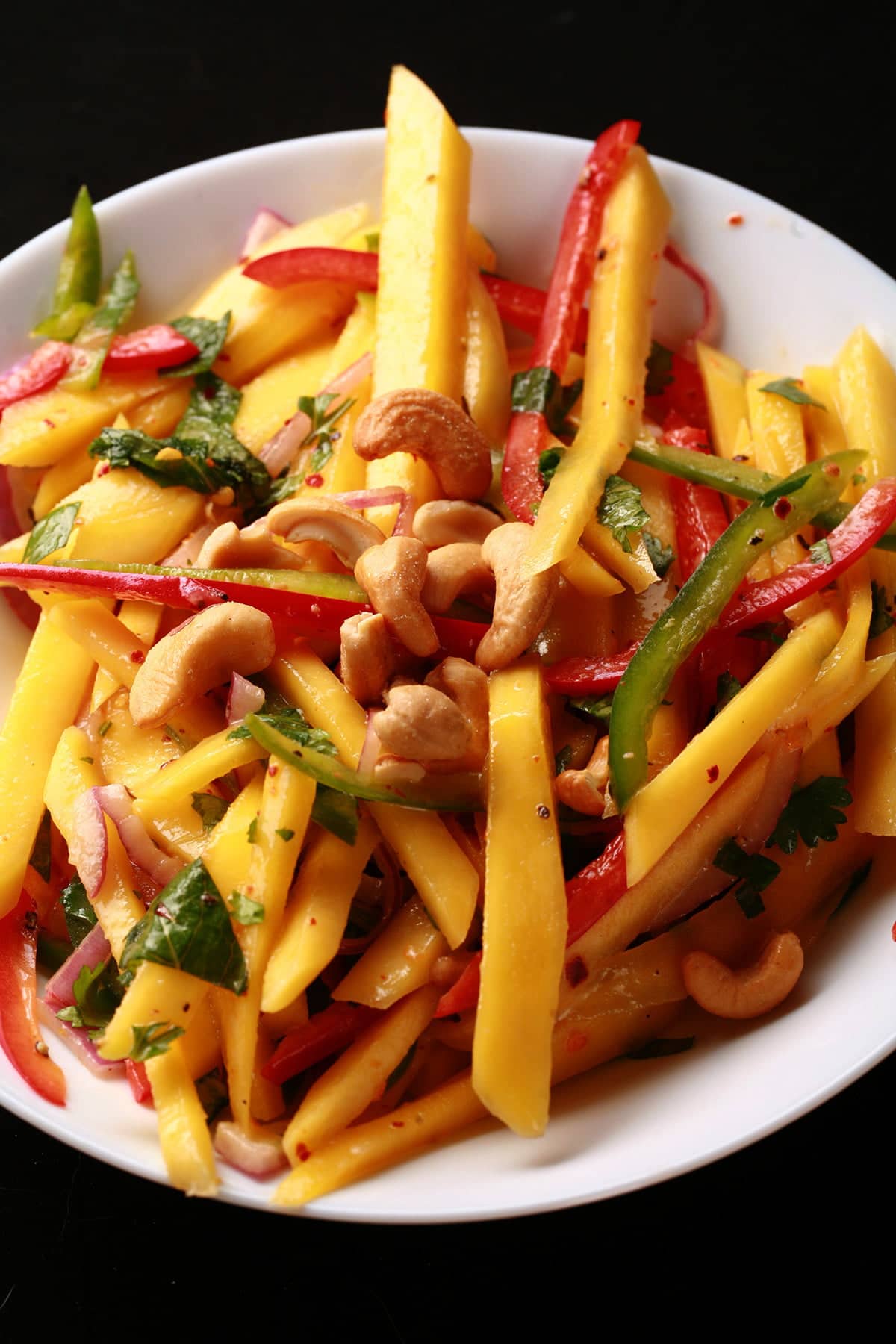 A close up view of Mango Salad - thin sticks of mango, red pepper, green pepper, red onions, and cashews. It's dressed with lime juice and pieces of cilatro and mint.