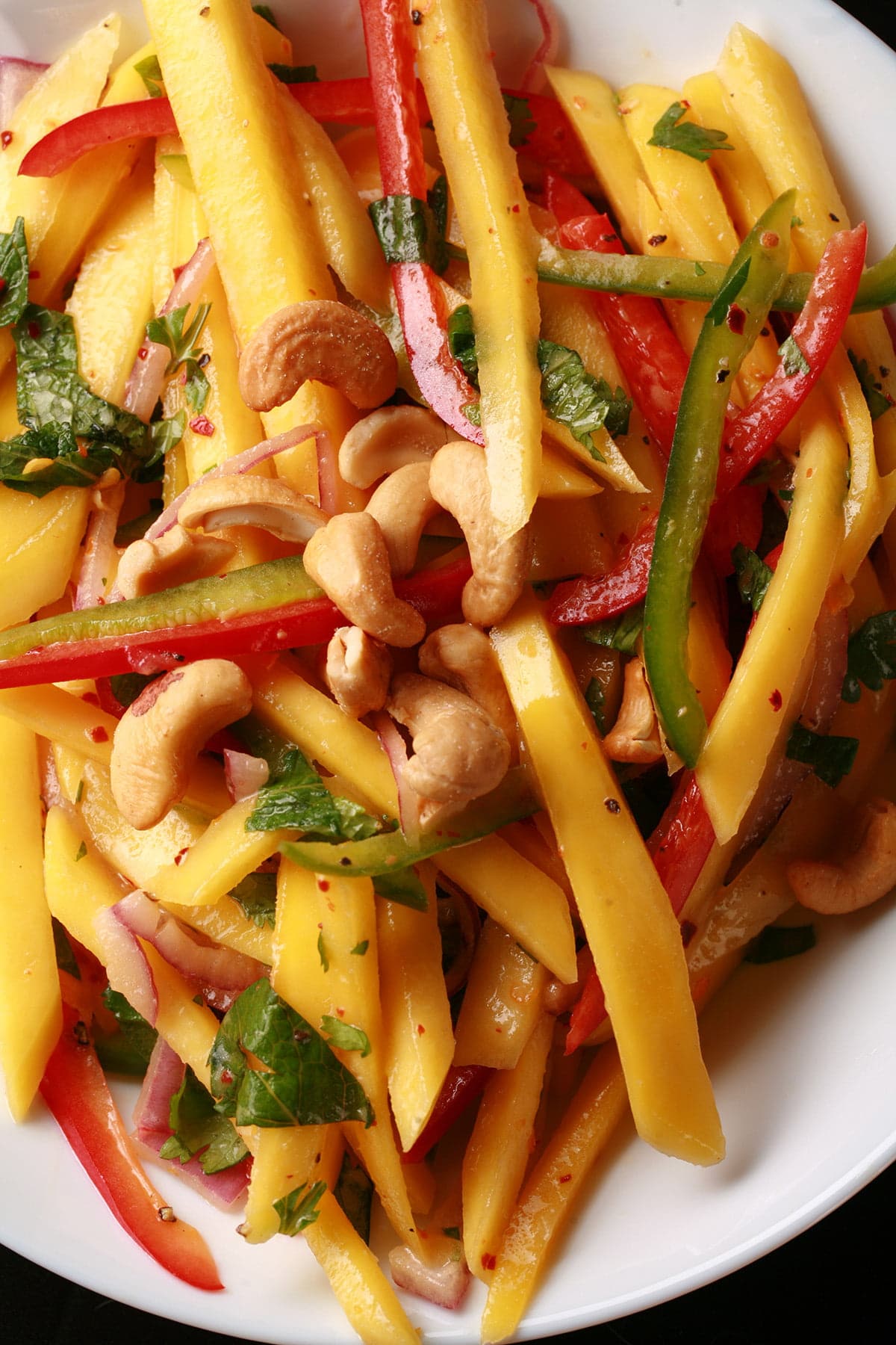 A close up view of Mango Salad - thin sticks of mango, red pepper, green pepper, red onions, and cashews. It's dressed with lime juice and pieces of cilatro and mint.