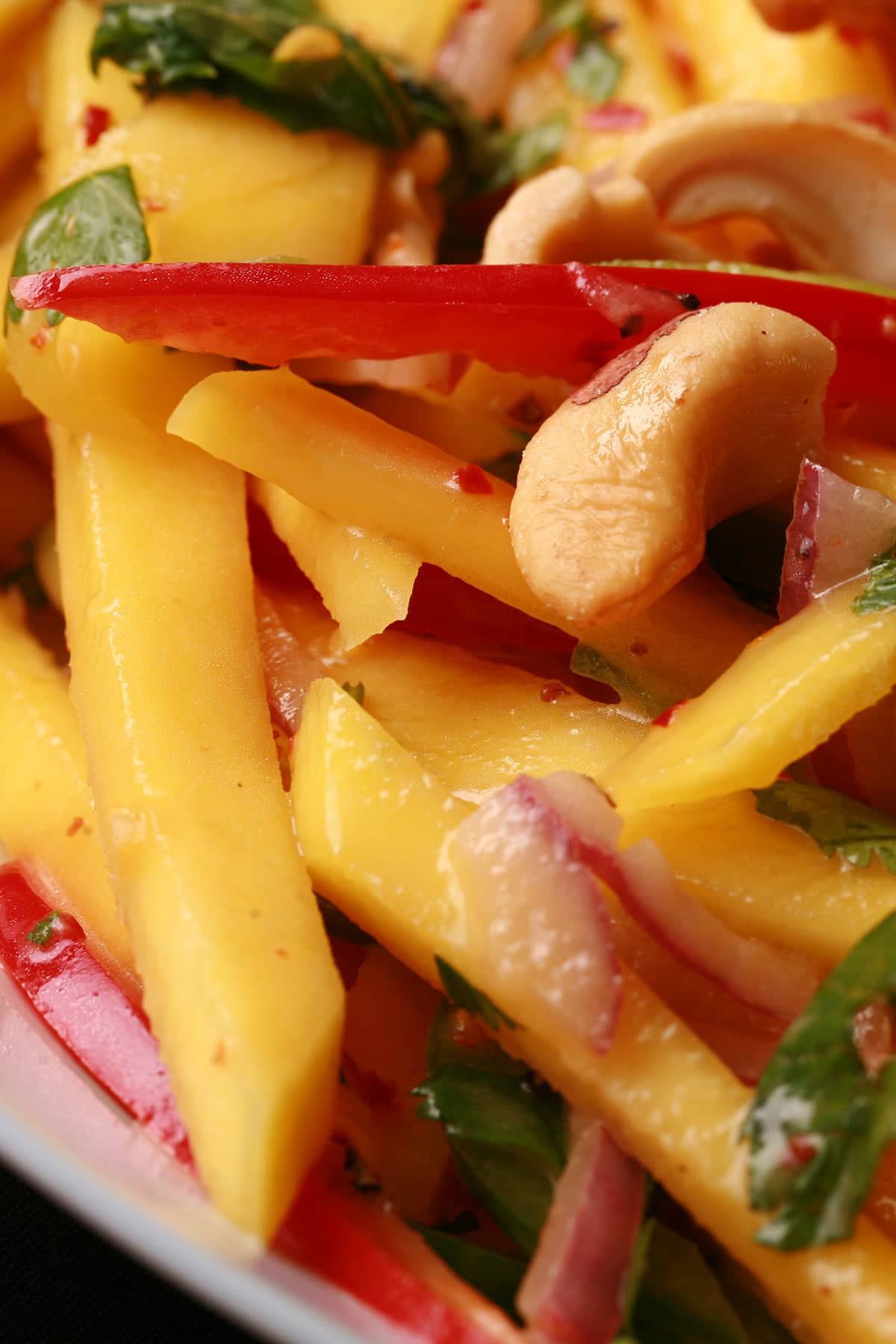 A close up view of Mango Salad - thin sticks of mango, red pepper, green pepper, red onions, and cashews. It's dressed with lime juice and pieces of cilatro and mint.