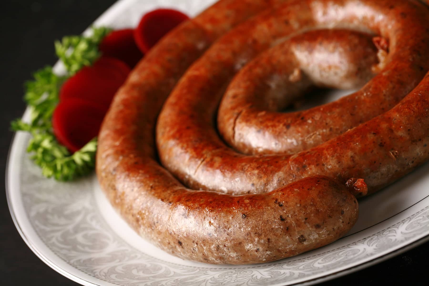 A large coil of potato sausage on a white plate. It's garnished with slices of beet pickles and some parsley.