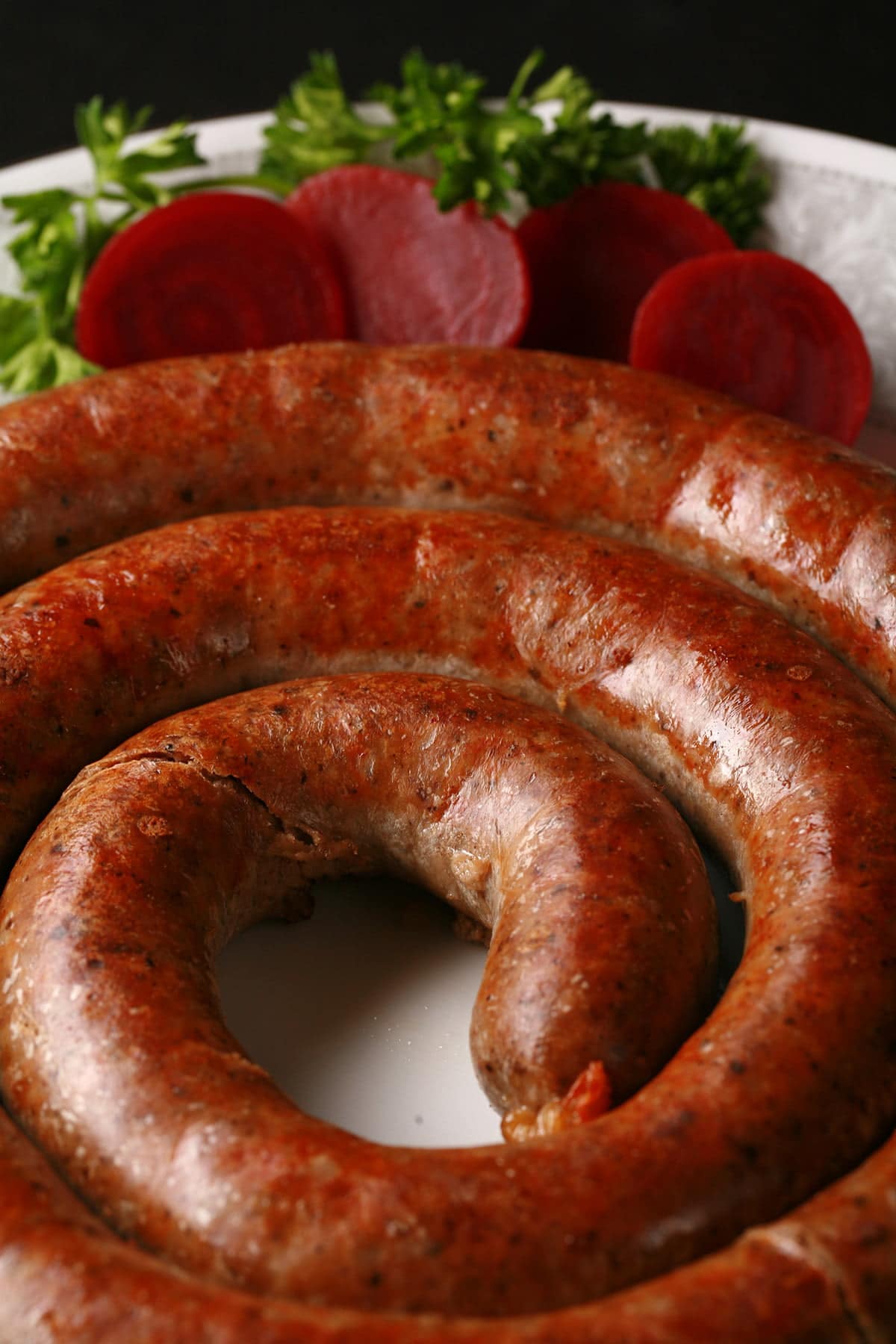 A large coil of potato sausage on a white plate. It's garnished with slices of beet pickles and some parsley.