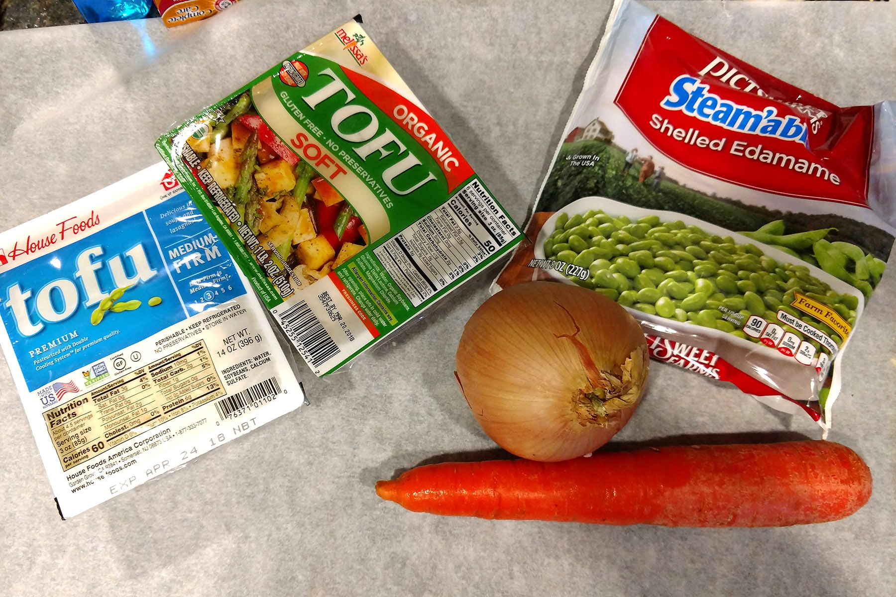 A carrot, an onion, a bag of frozen edamame, and 2 packs of tofu on a countertop.