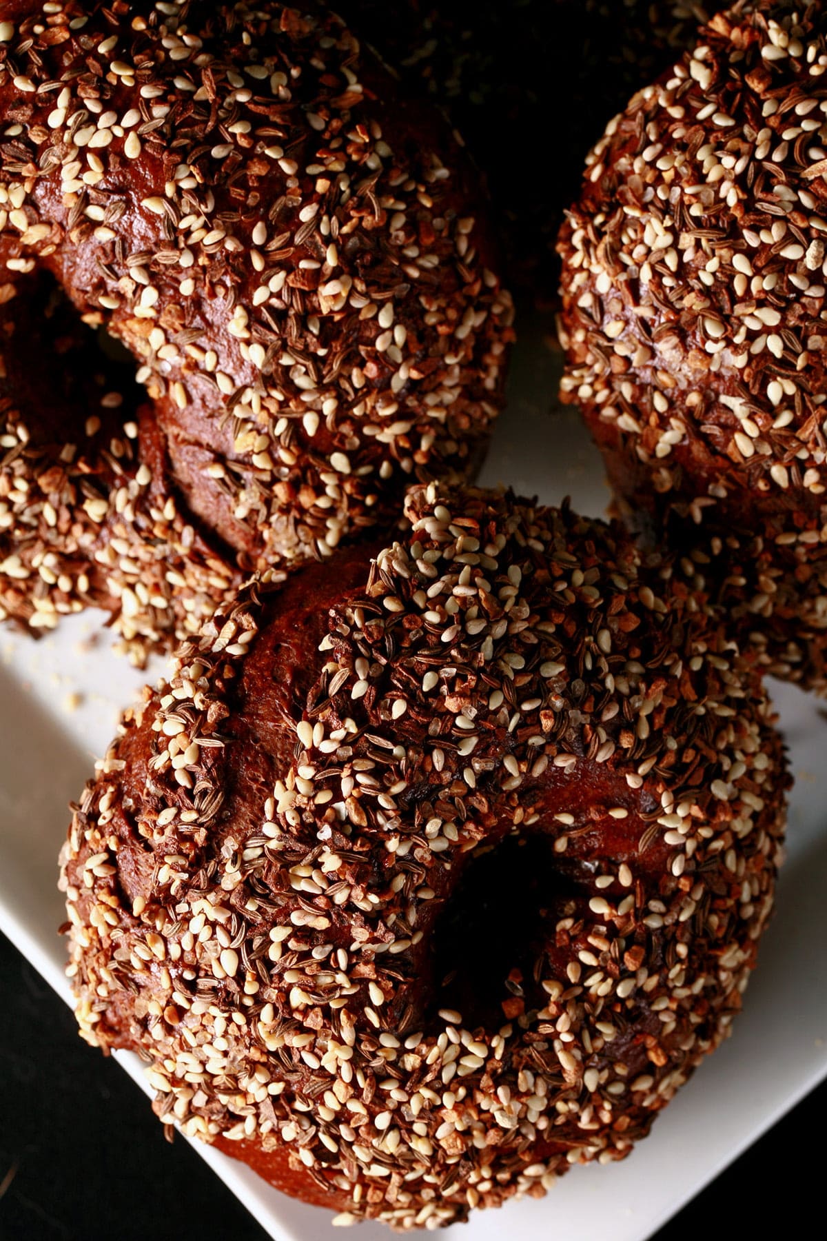 Several "Everything" style Pumpernickel Bagels on a square white plate.