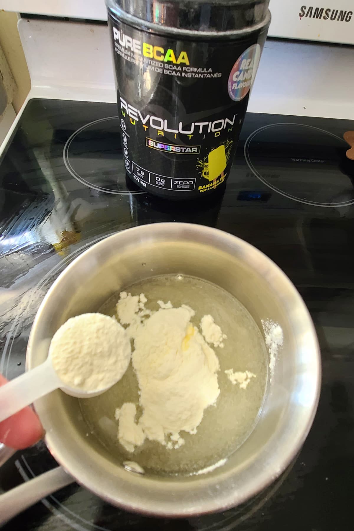 Yellow BCAA powder being added to a small pot of water. A container of Revolution Nutrition's Banana BCAA powder is in the background.