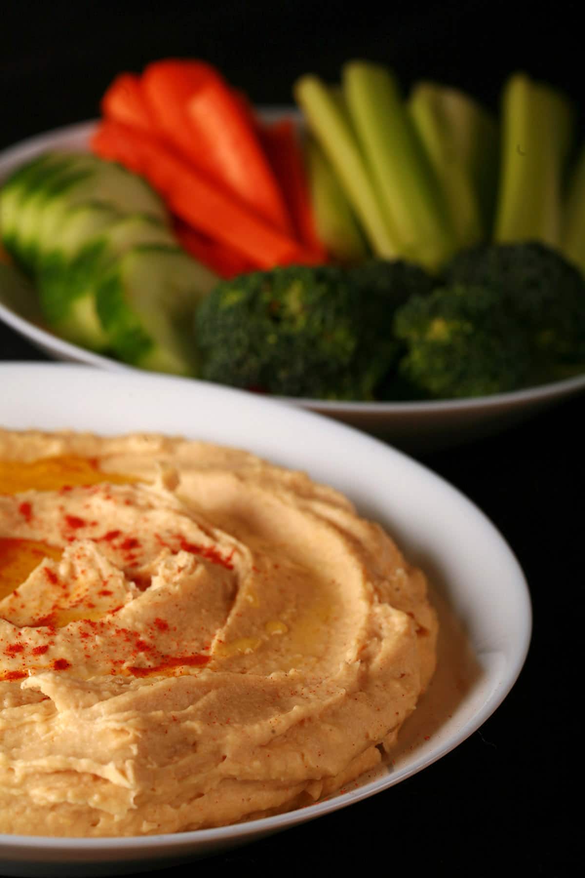 A white bowl of smooth hummus.  There is olive oil and a sprinkling of paprika on top, and a bowl of vegetable dippers next to it.