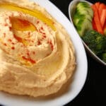 A white bowl of smooth hummus. There is olive oil and a sprinkling of paprika on top, and a bowl of vegetable dippers next to it.
